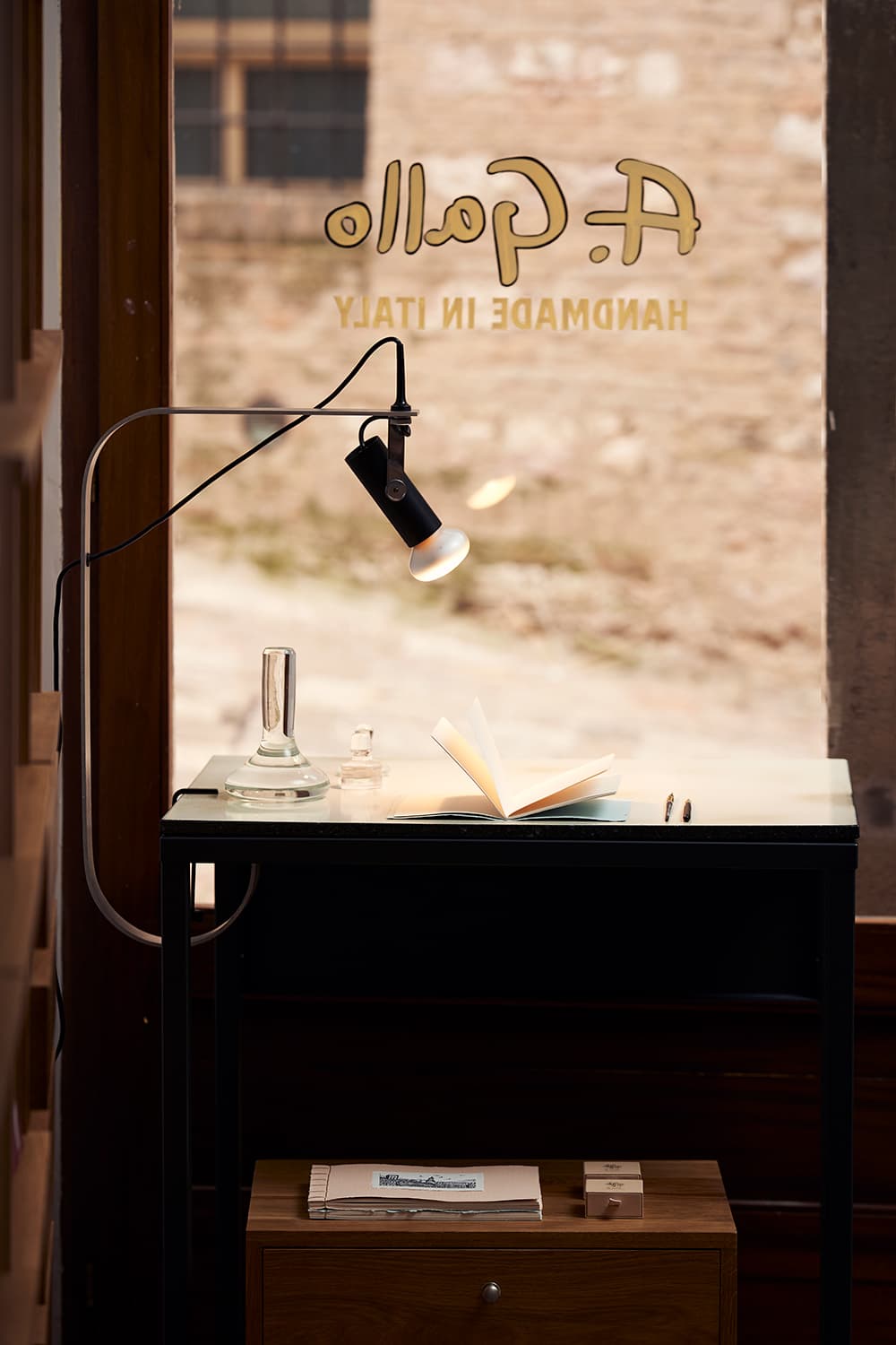 A desk with a lamp, open notebook, and glass bottle, set against a window.