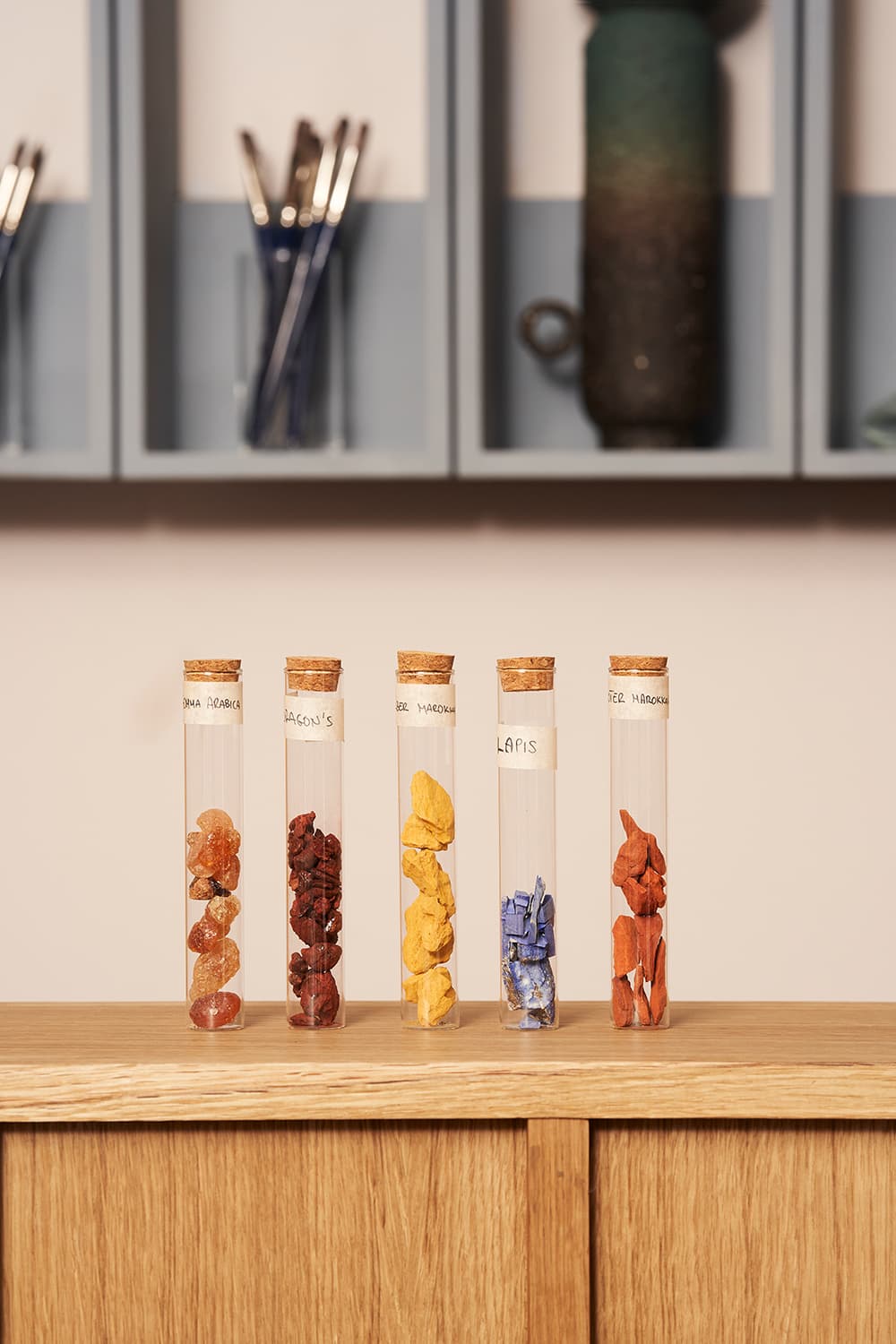Five glass vials with cork lids containing colorful stones are displayed on a wooden surface, with shelves and brushes in the background.