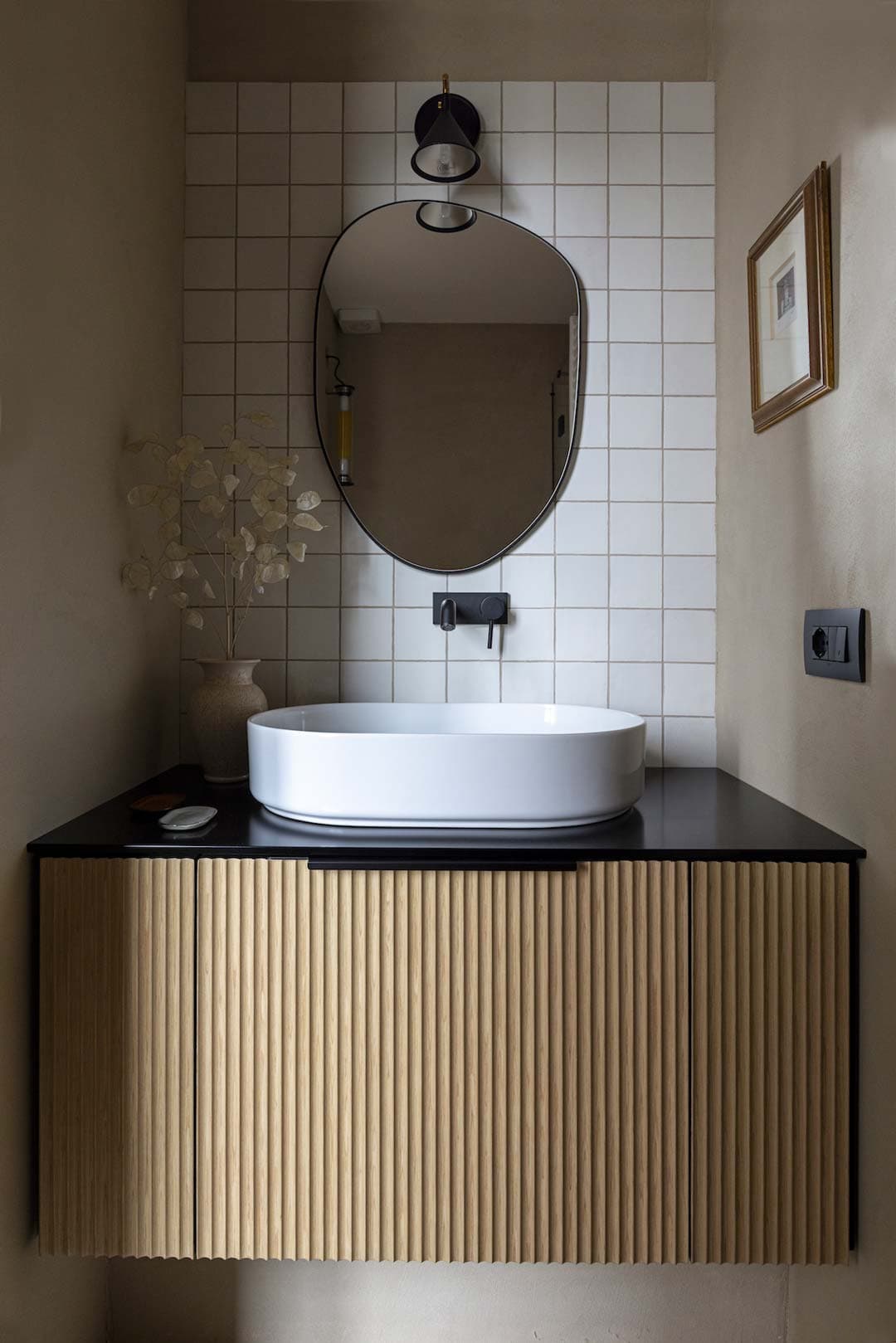 Modern bathroom with oval mirror, white sink on a black countertop, wood-textured cabinet, and tiled wall. Minimalist decor with a wall light.