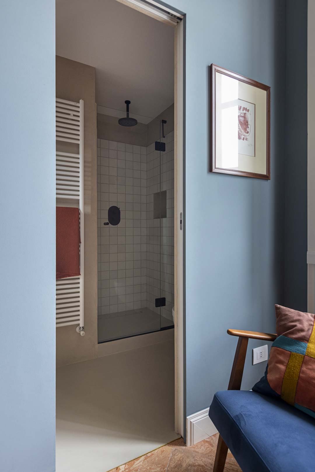 A modern bathroom with a glass shower and white tiles. A blue chair and framed picture are visible in the foreground.
