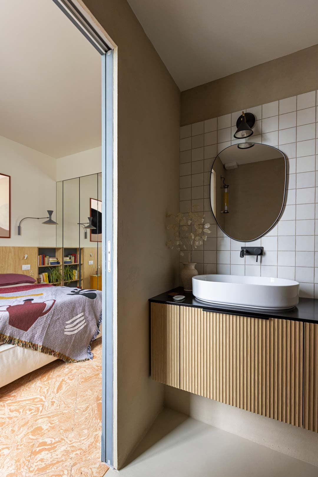 Modern bathroom with a round mirror, wall-mounted faucet, and vessel sink. Adjacent bedroom features a patterned bedspread and bookshelves.