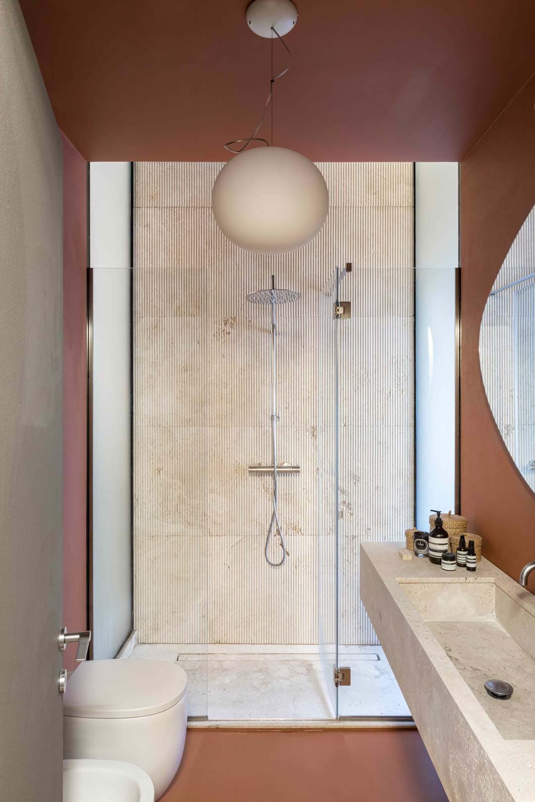 Modern bathroom with a glass shower, round white pendant light, beige countertop with toiletries, and terracotta walls and floor.