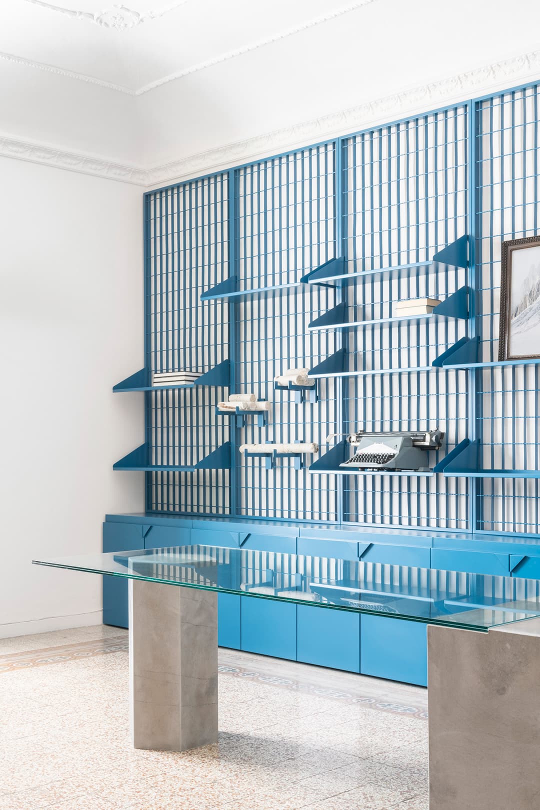 Modern office with a glass table, blue shelving on the wall, and a typewriter. Bright and minimalistic design with white walls and terrazzo floor.