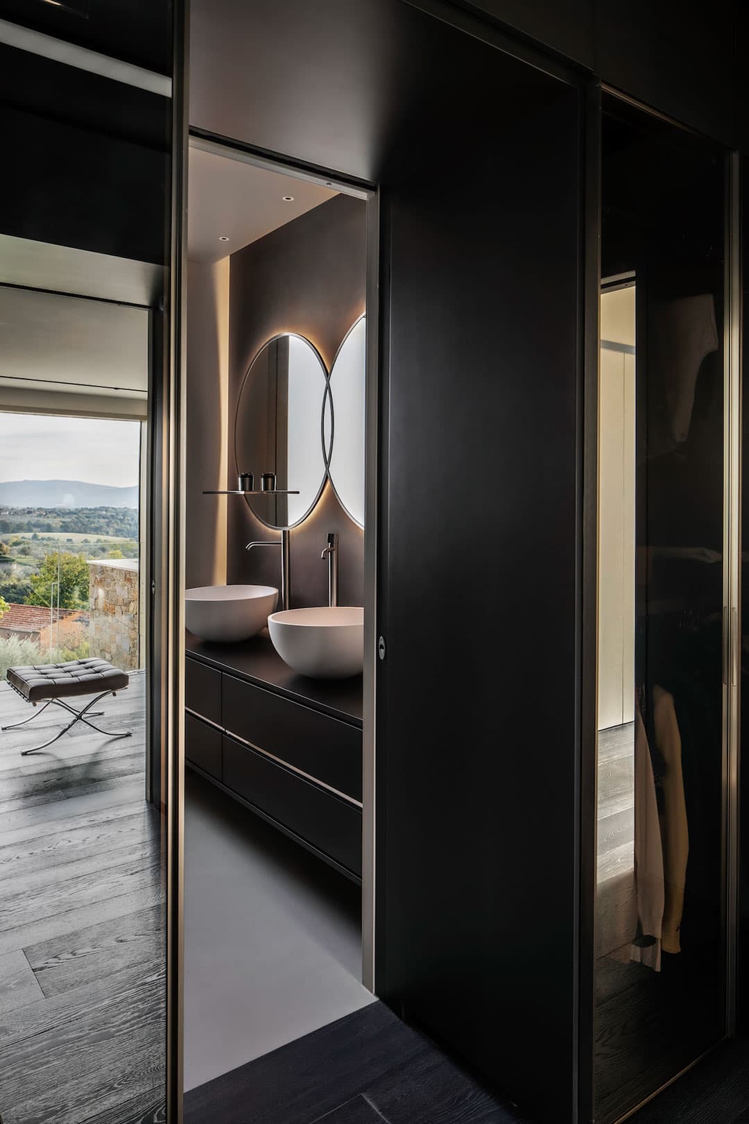 Modern bathroom with two round mirrors, vessel sinks, and dark cabinetry. Sliding door partially open, revealing a scenic view outside.