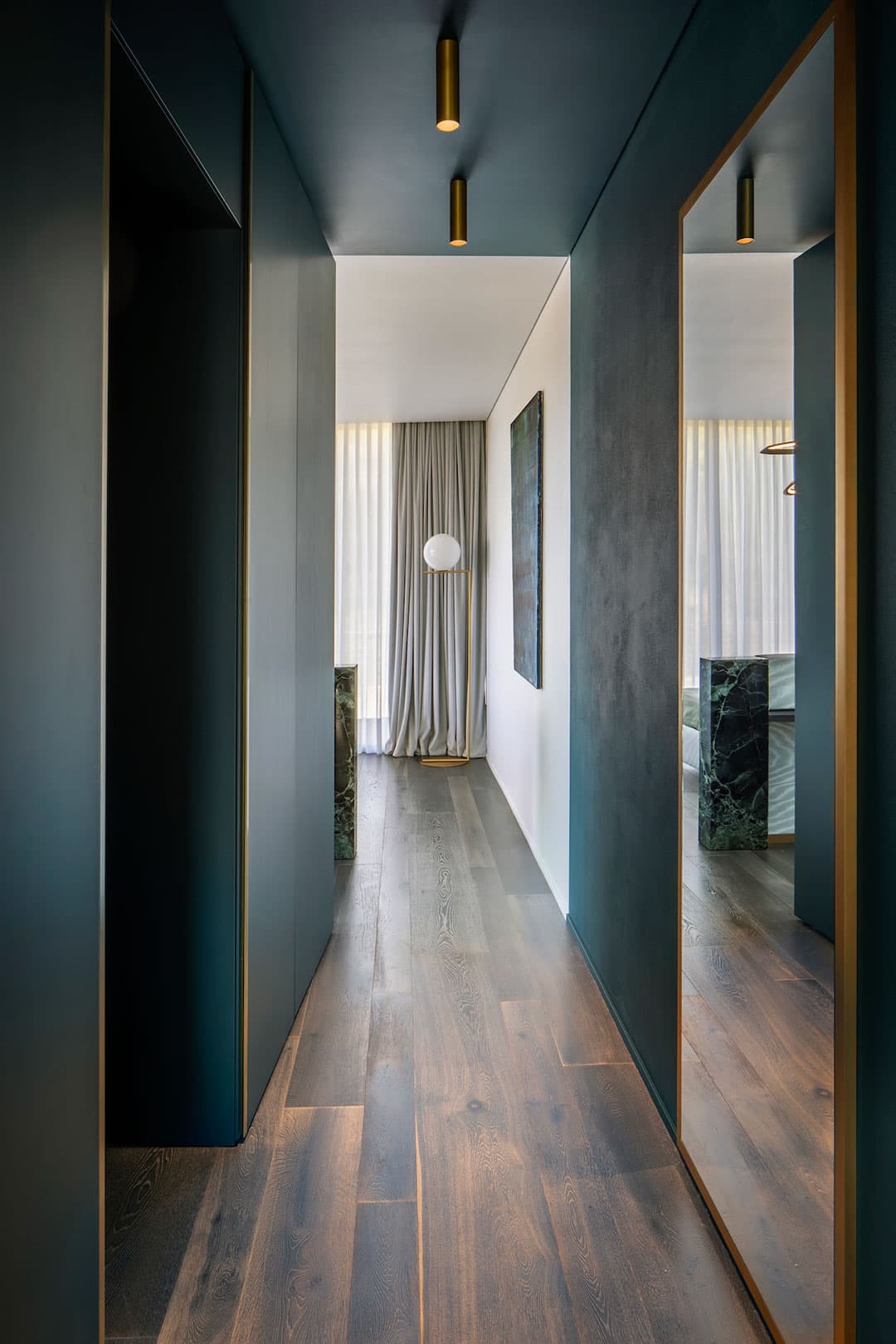 A modern, narrow hallway with dark walls, wooden flooring, a large mirror, ceiling lights, and a window with curtains in the background.