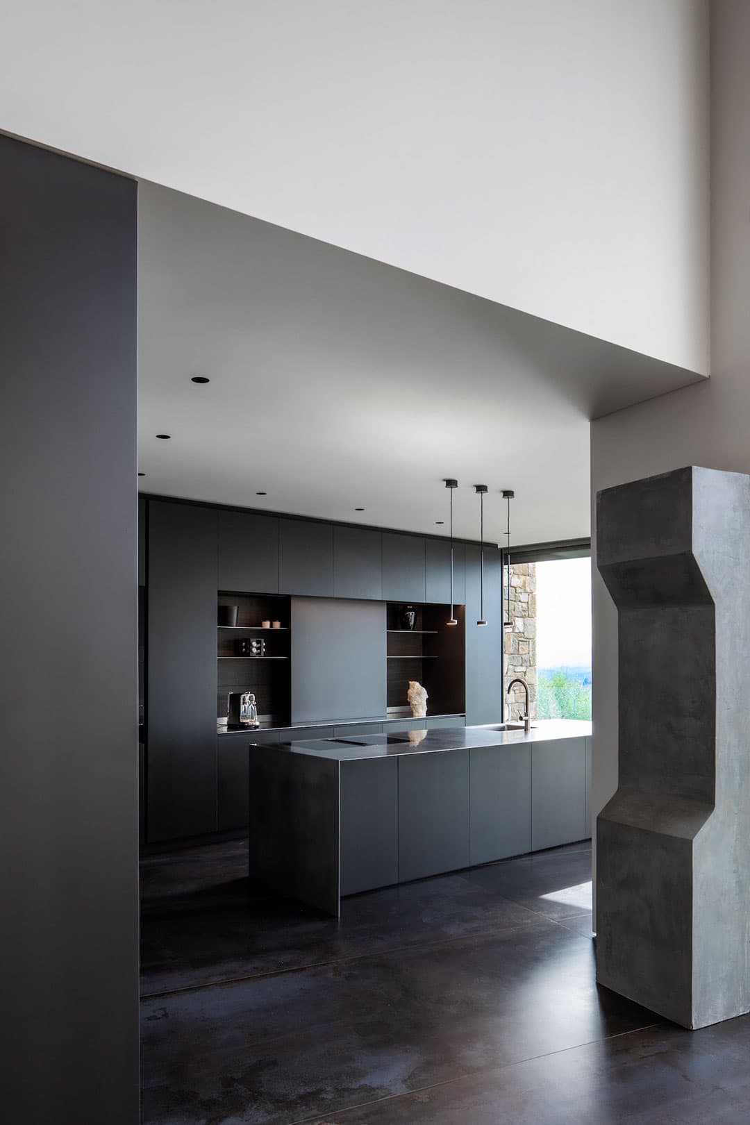 Modern kitchen with dark cabinetry, a large island, pendant lights, and minimalist decor. A tall, geometric structure is visible on the right.