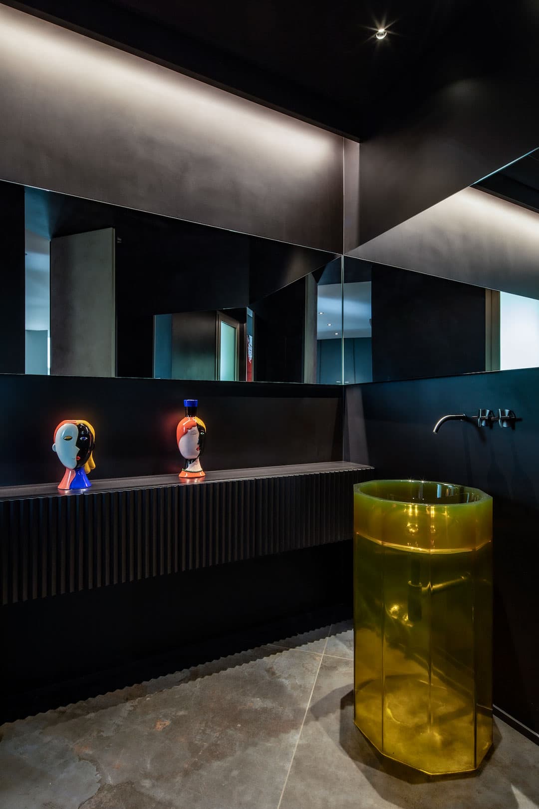 Modern bathroom with dark walls, large mirror, and a unique yellow translucent pedestal sink. Two abstract bust sculptures on the counter.