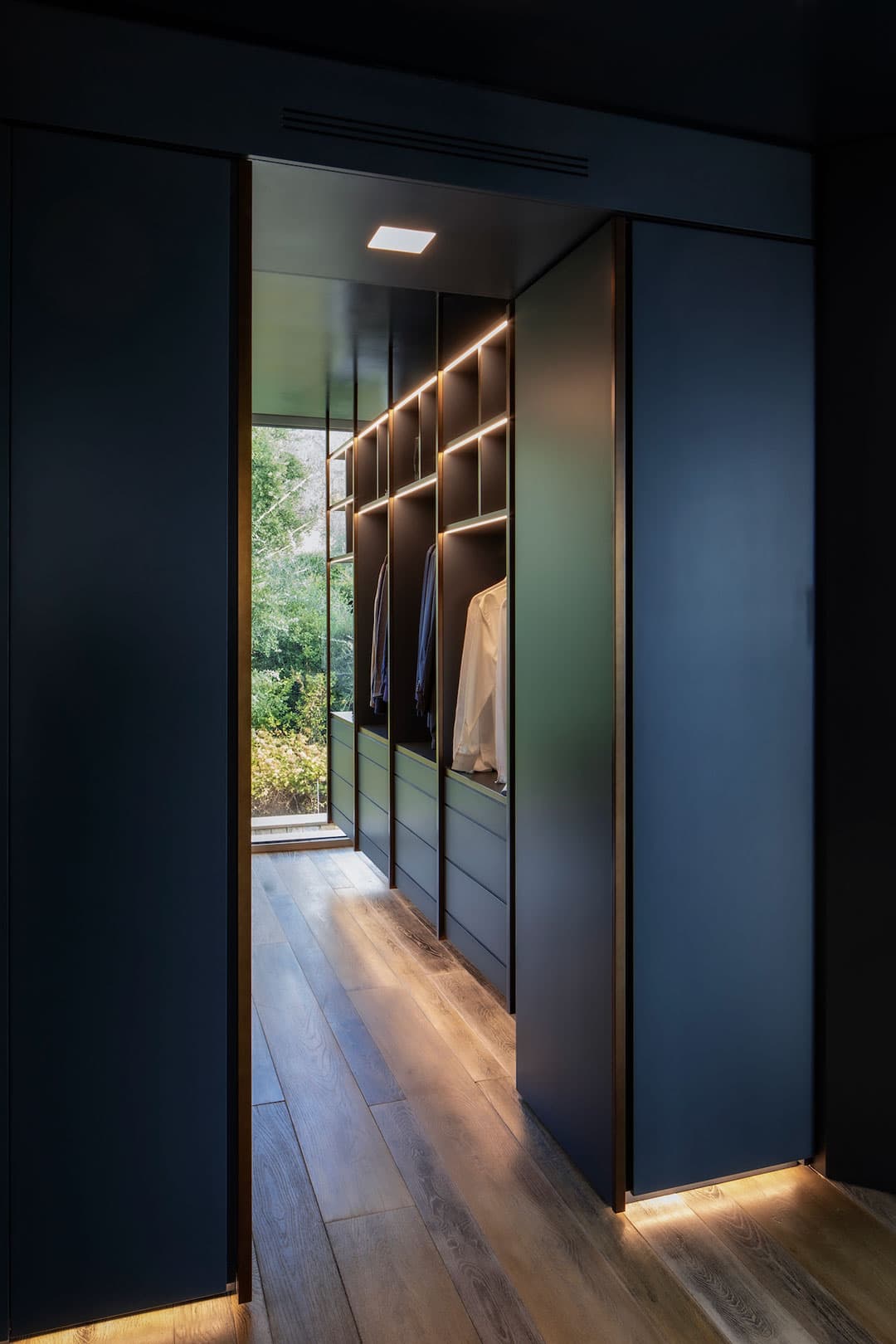 Modern walk-in closet with dark cabinets, wooden floor, and clothes hanging. Large window offers a view of greenery outside.