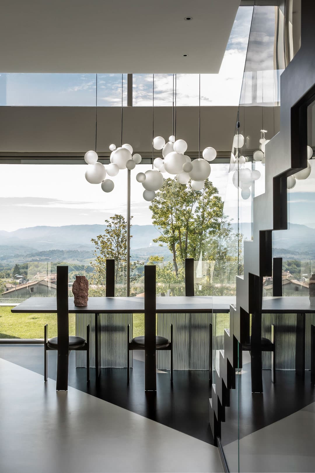 Modern dining area with a sleek black table, unique chairs, and a cluster of white pendant lights. Large windows offer a scenic view of mountains.