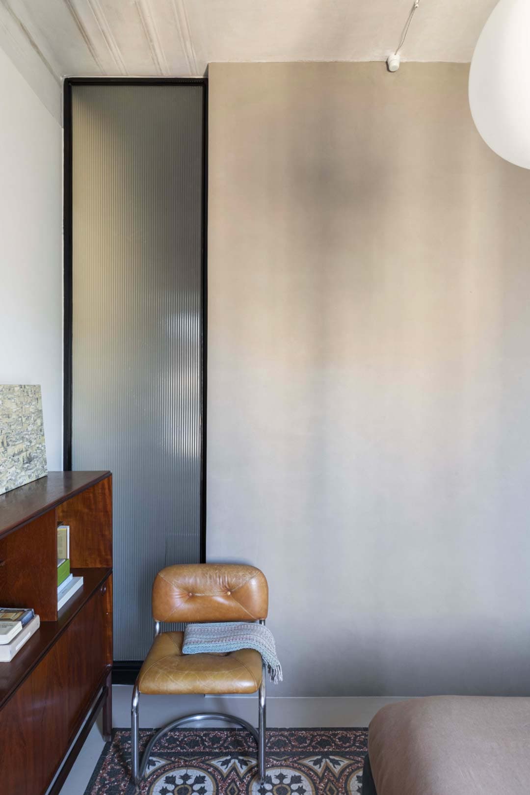 A minimalist room with a brown leather chair, wooden cabinet and patterned floor tiles. A frosted glass panel divides the space.