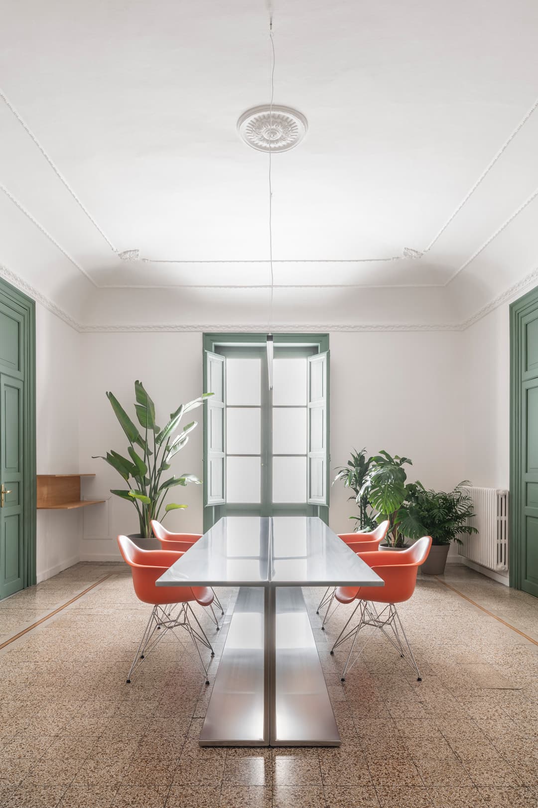 A minimalist room with a long table, orange chairs and large green plants.