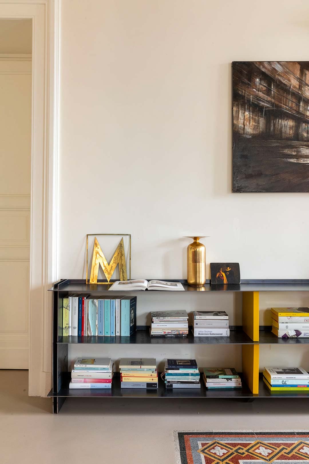 Modern interior with a black shelf holding books, a gold vase, and framed letter 'M'.