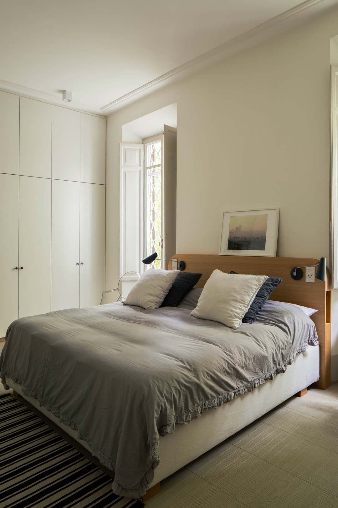 Cozy bedroom with a large bed, gray bedding, and pillows. A painting rests on the headboard. Wardrobe and window with natural light.