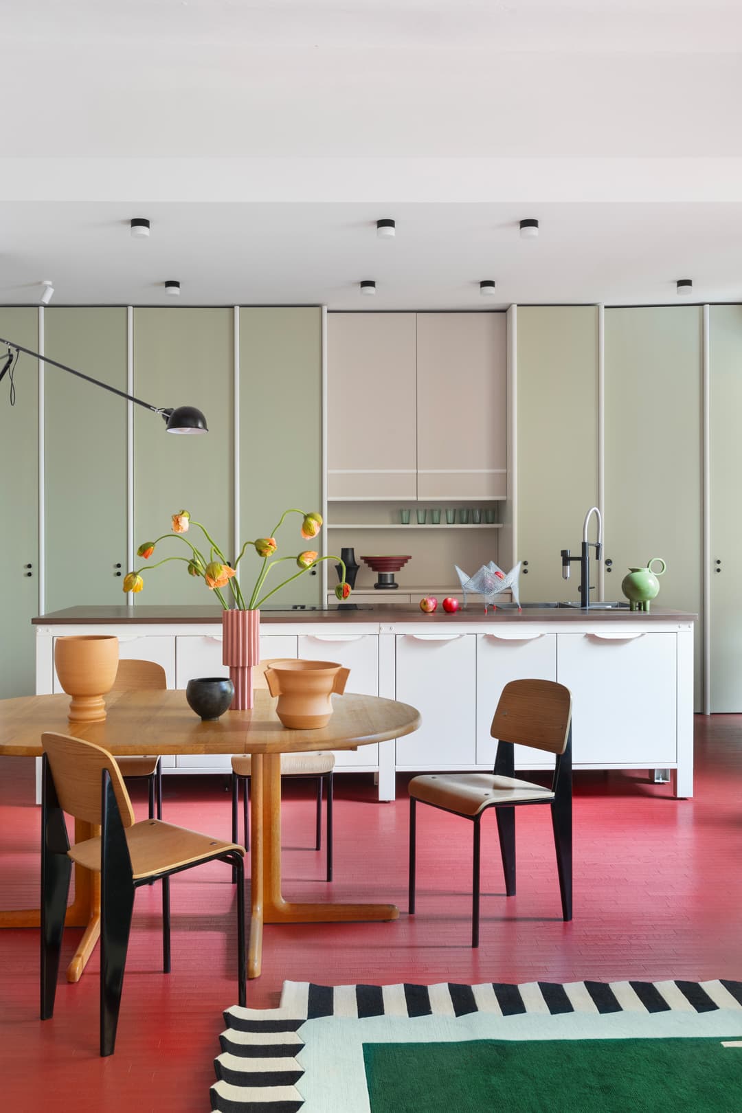 Modern kitchen with red flooring, round wooden table, chairs, white cabinets, and a vase with flowers. A rug in foreground.