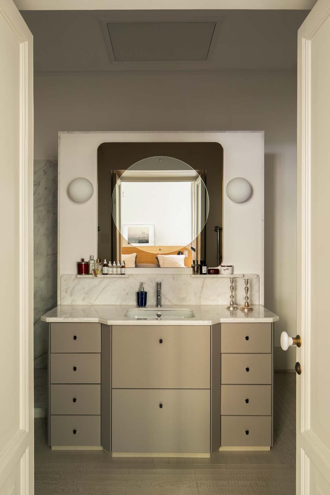 Modern bathroom with a round mirror, marble countertop, and sleek cabinetry. Reflections show a bed in the adjacent room.