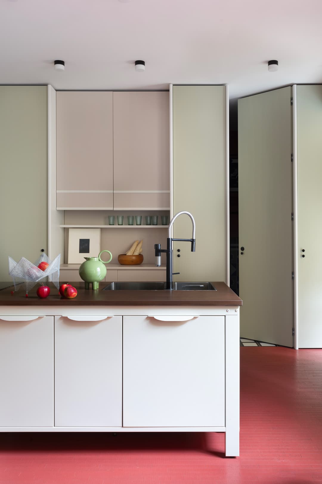 Modern kitchen with a large island, black sink, and faucet. Green teapot on the counter. Light cabinets and red floor.
