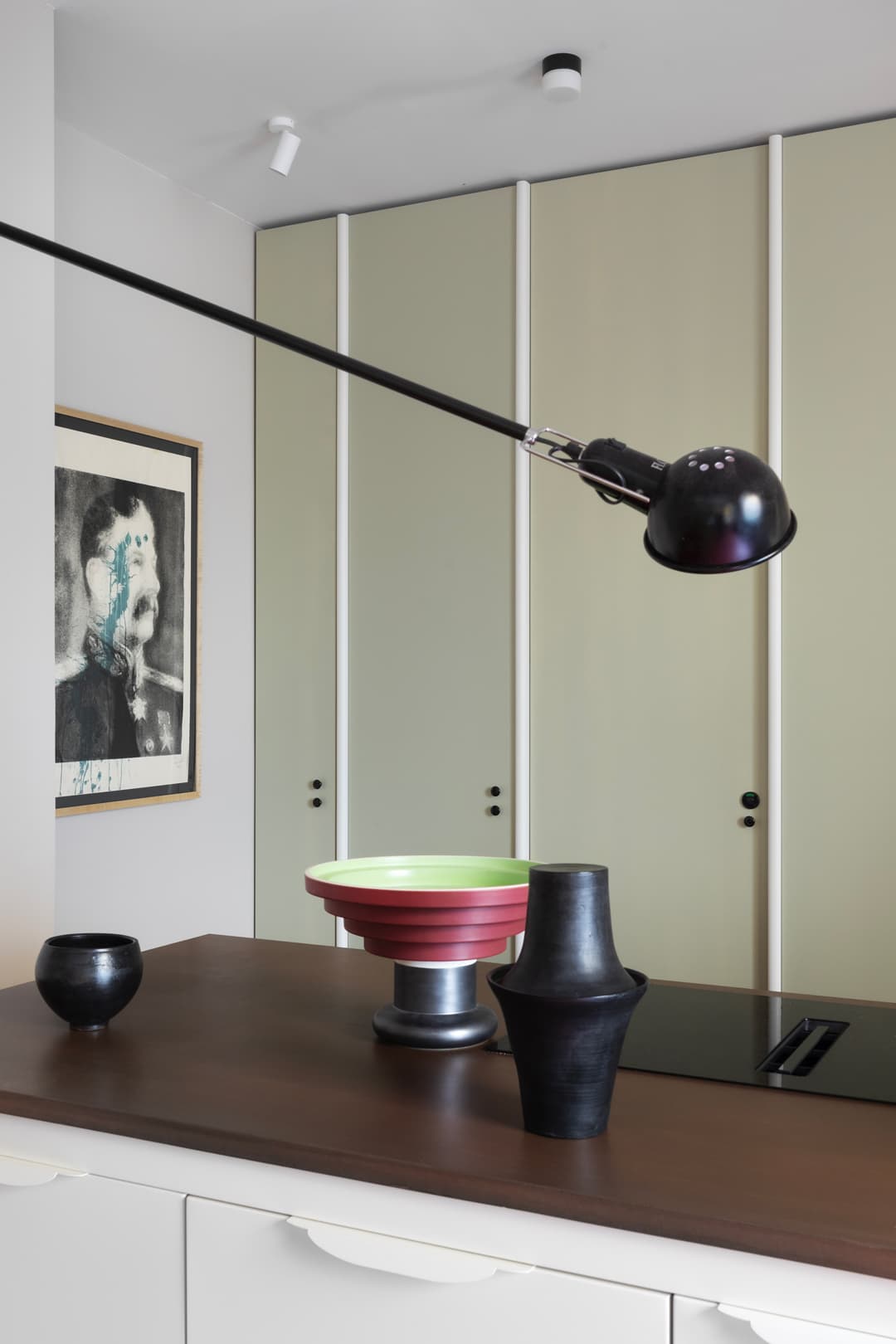 Modern kitchen interior with a dark countertop, decorative vases, wall art, and sleek cabinetry. A black overhead lamp adds a contemporary touch.