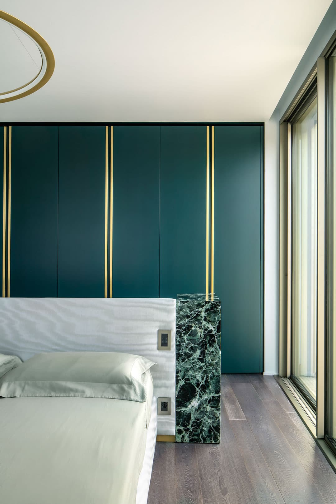 Modern bedroom with a green and gold accent wall, a marble-topped headboard, and large windows.