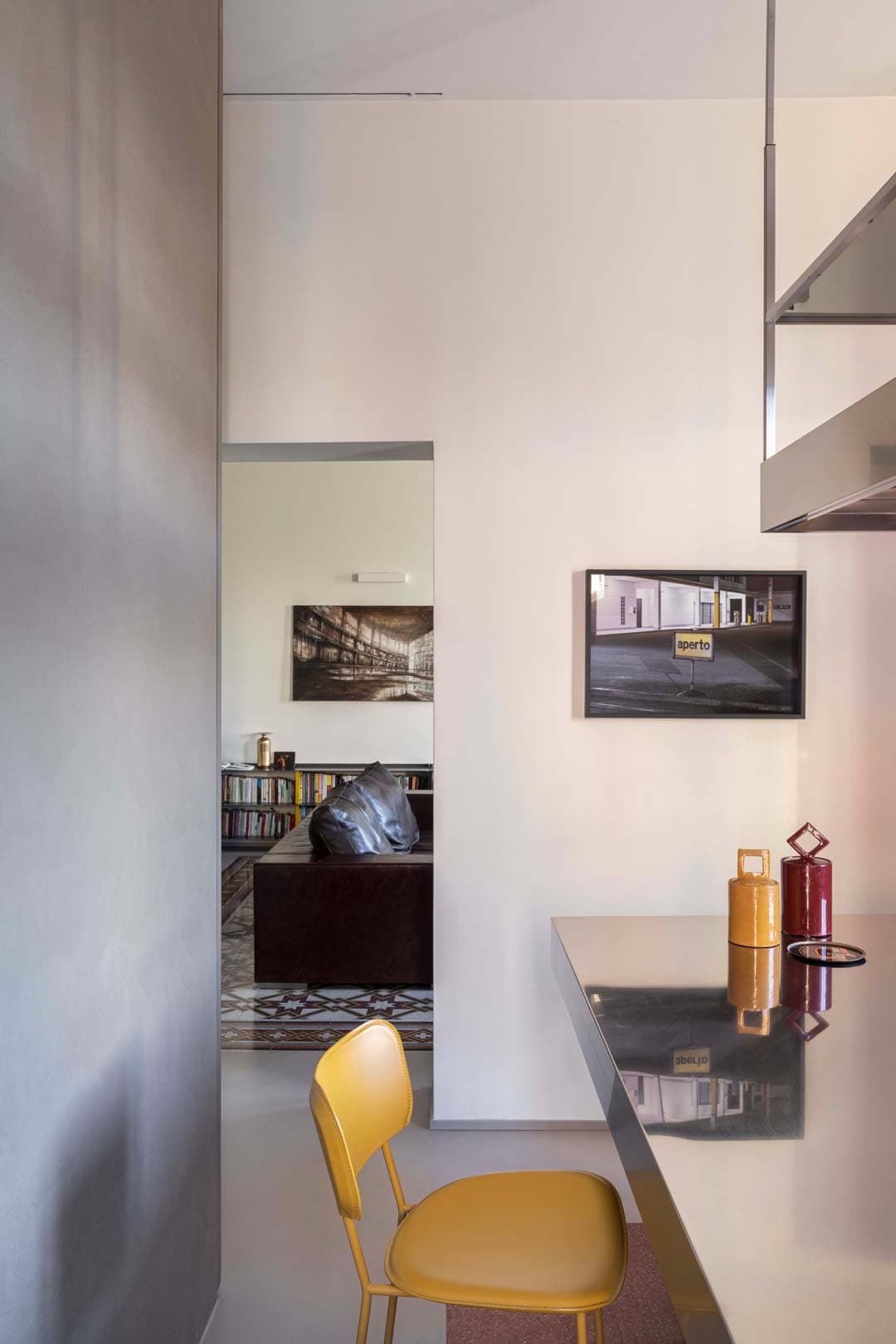 Modern interior with a yellow chair by a sleek countertop and a glimpse into a cozy living area with bookshelves and a sofa.