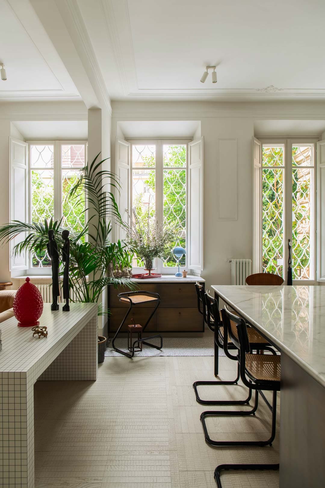 Modern dining area with large windows, plants, and a large marble island surrounded by black chairs. Decor accent the space.