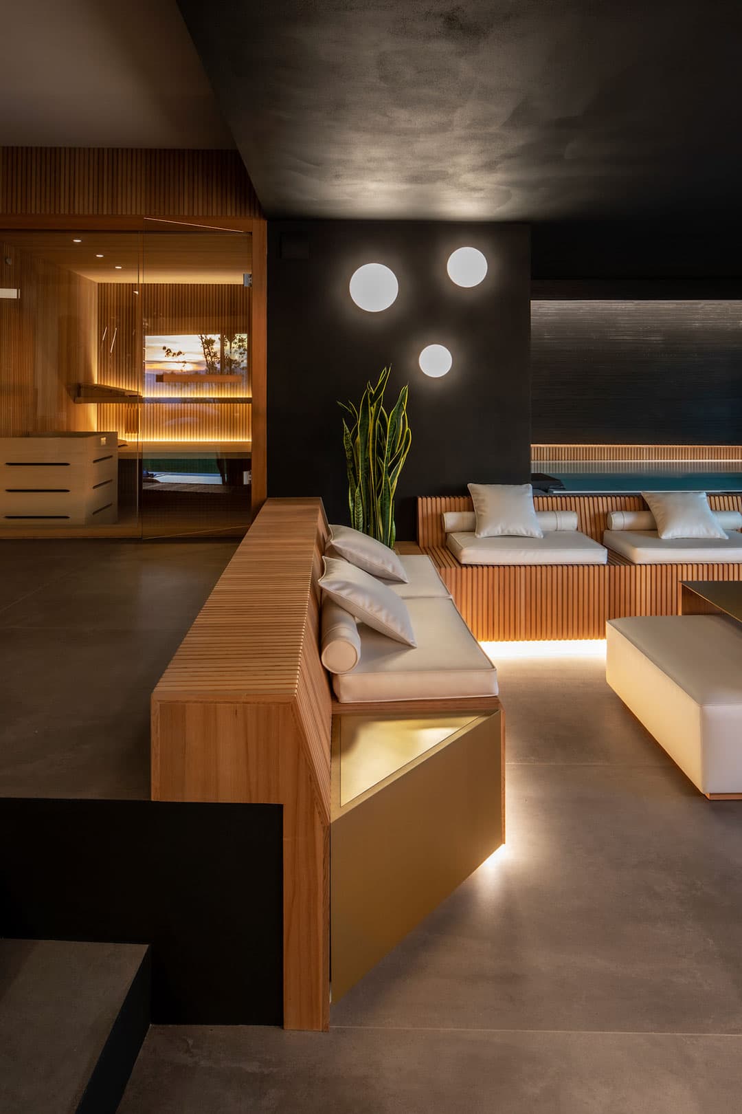 Modern lounge with wooden seating, ambient lighting, and wall-mounted circular lights. Cushions on benches and a potted plant in the corner.