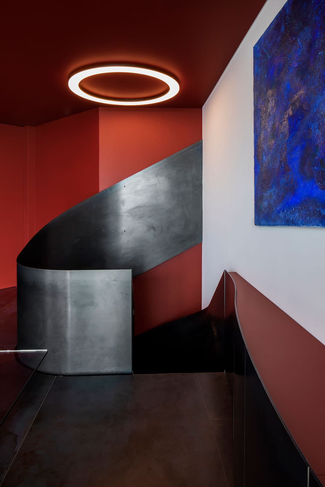 Modern interior with a curved metal staircase, red walls, a circular ceiling light, and a blue abstract painting on a white wall.