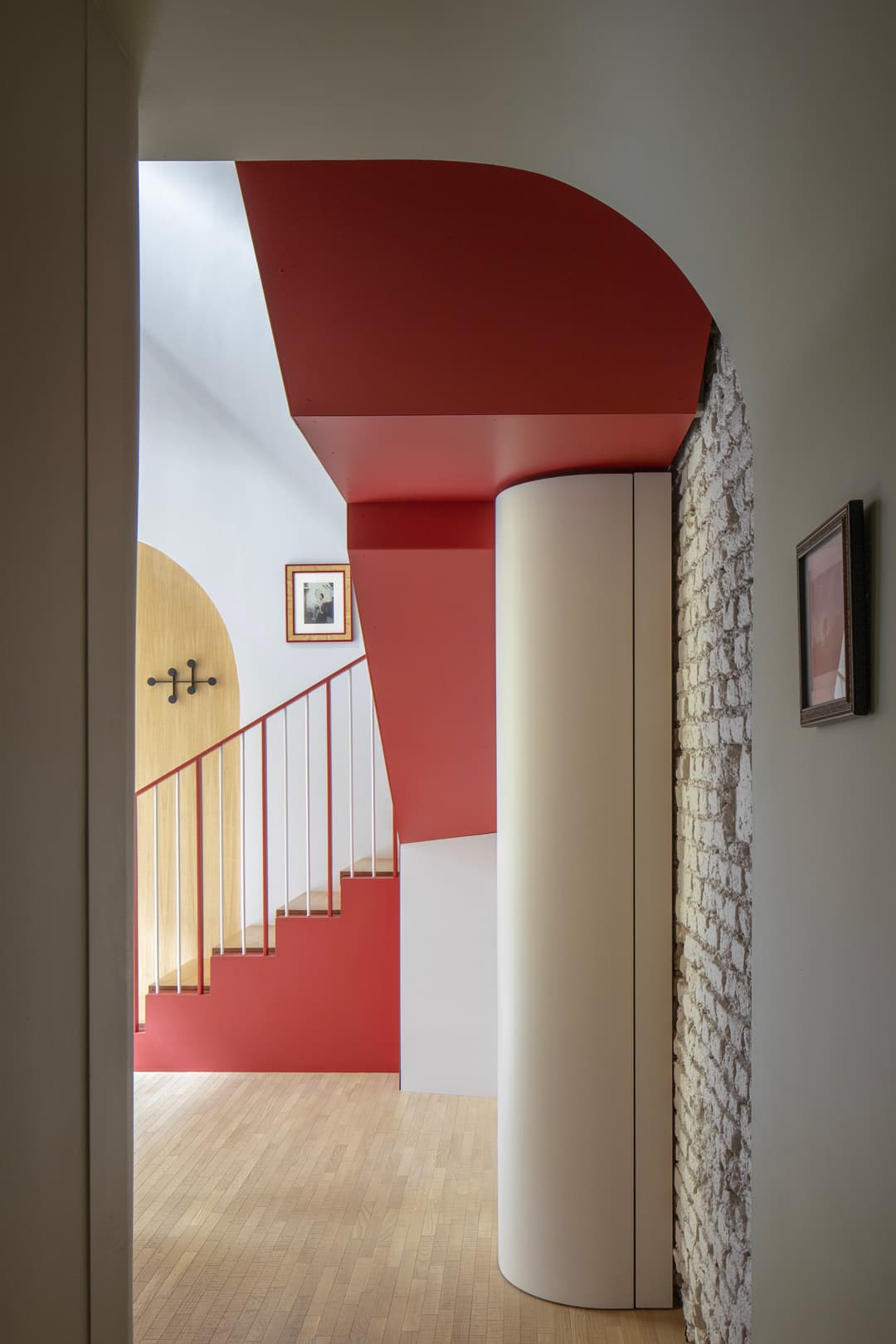 Modern interior with red accents, a curved white pillar, brick wall, wooden staircase, and framed artwork.