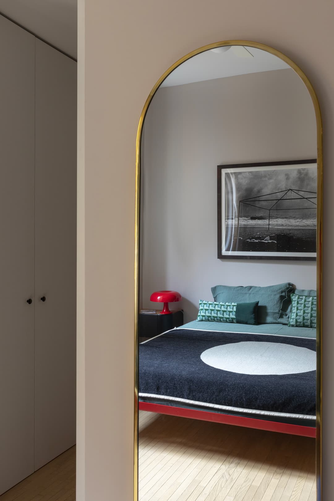 Arched mirror reflecting a modern bedroom with a bed, red lamp, and framed picture. Wooden floor and closed white closet door visible.
