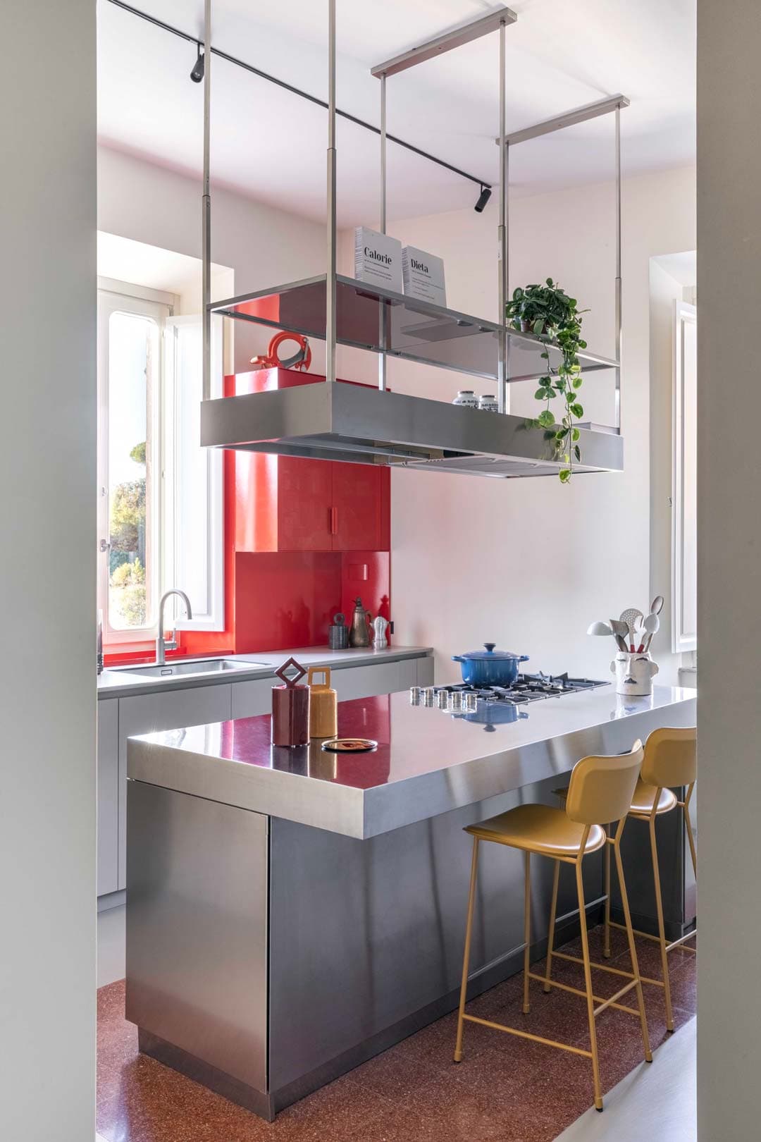 Modern kitchen with a sleek metal island, yellow bar stools, hanging shelves with plants, and a window with natural light.
