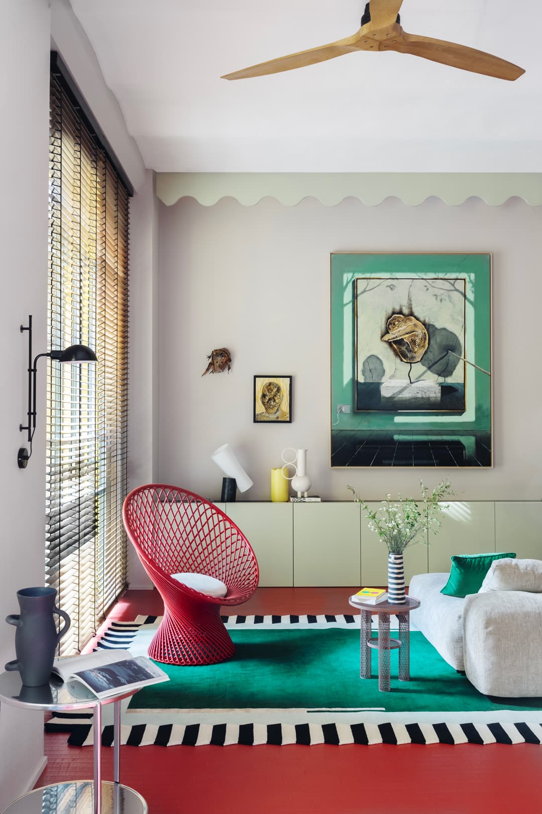 Modern living room with a red chair, green rug, and abstract art. Features a ceiling fan and large window with blinds.