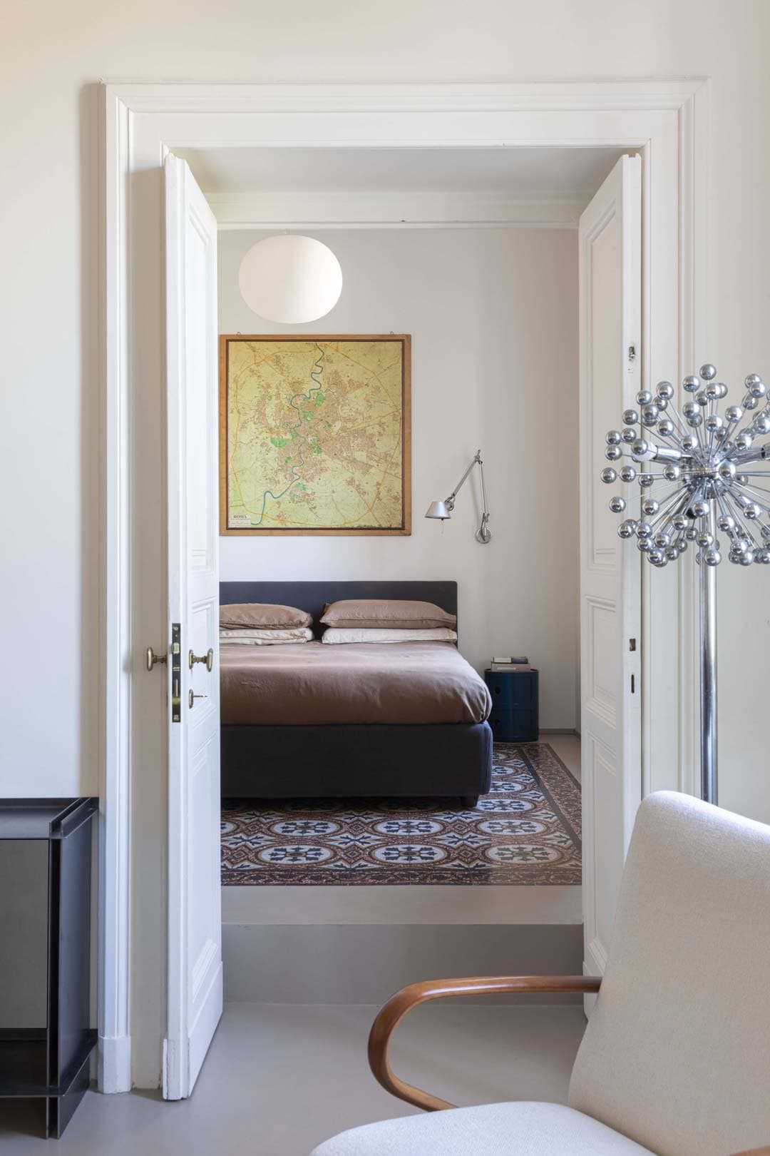 A cozy bedroom with a bed, map artwork on the wall, and a patterned rug, viewed through open white double doors.