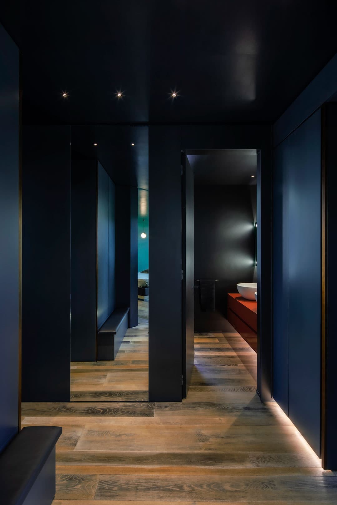 Modern hallway with dark walls, wooden floors, recessed lighting, and a glimpse of a bathroom featuring a vessel sink on a red vanity.