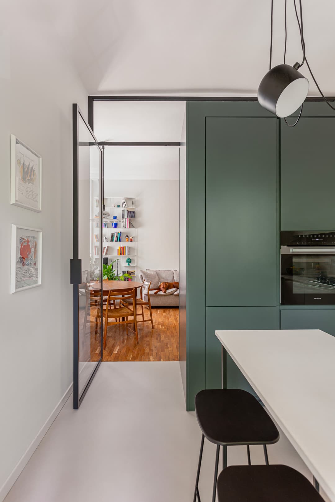 Modern kitchen with green cabinets, white table, and black stools. Glass door opens to a cozy living area with wooden flooring and bookshelves.