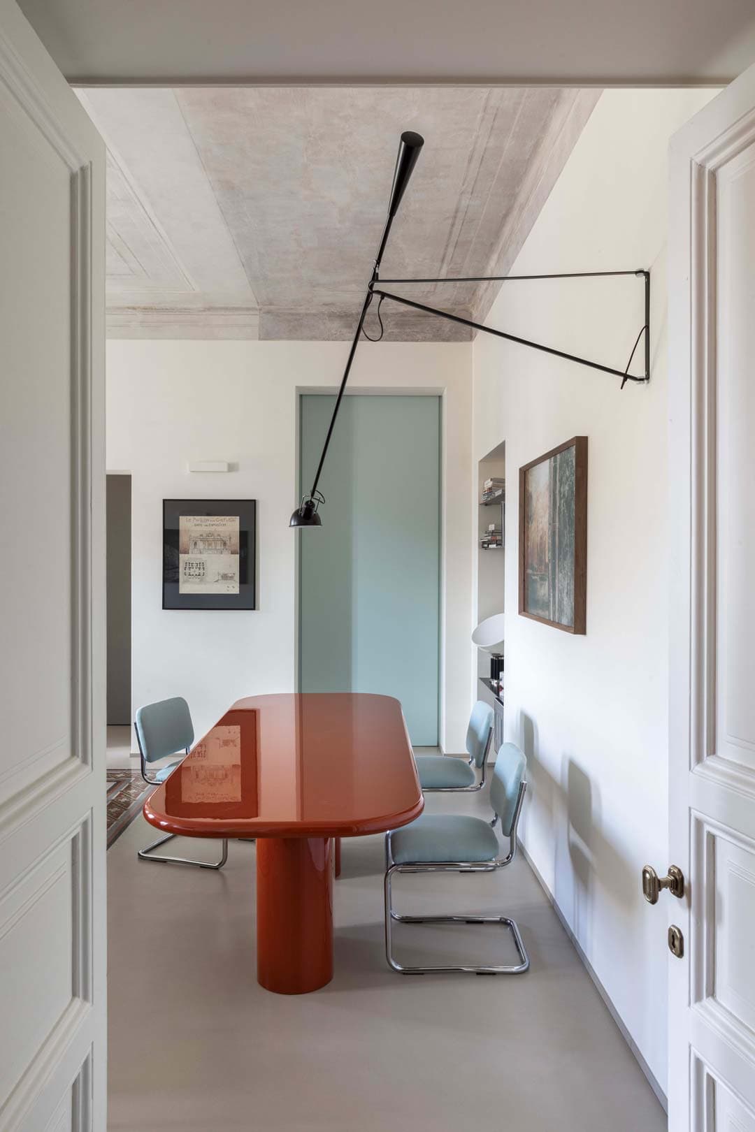 Minimalist room with a long red table, blue chairs, modern wall lamp, and framed art.