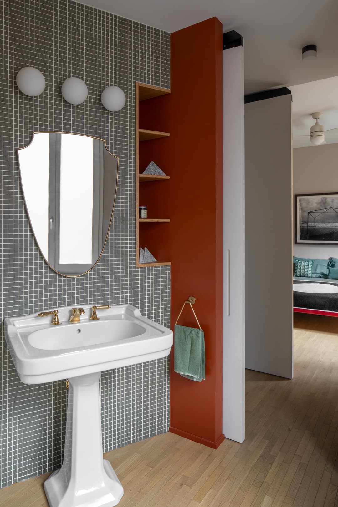 Bathroom with a pedestal sink, oval mirror, gray tile wall, and red accent wall with shelves. A towel hangs beside the sink. Doorway to another room.