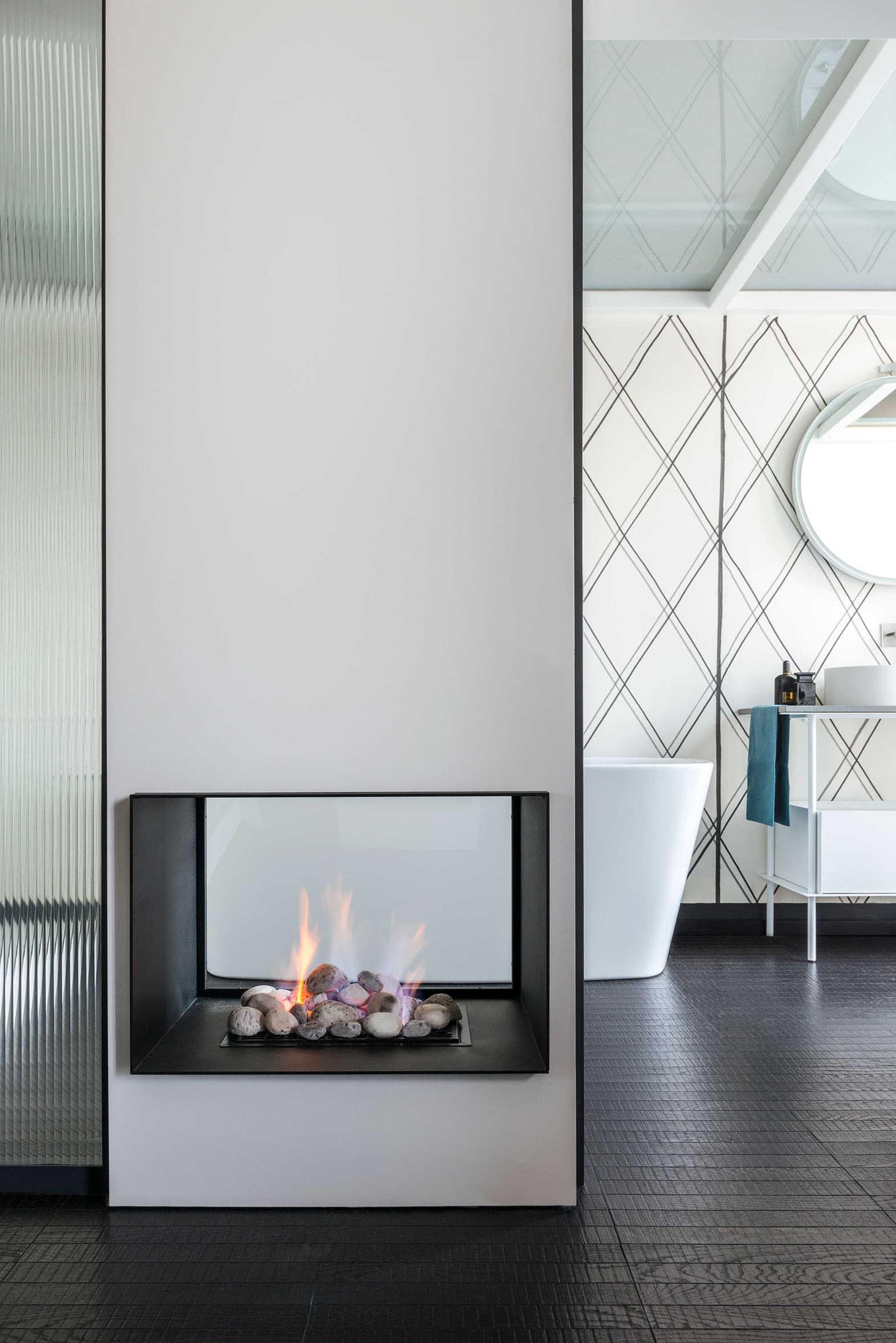 Modern bathroom with a sleek, built-in fireplace, freestanding bathtub, and a round mirror above a white vanity. Geometric wall pattern.