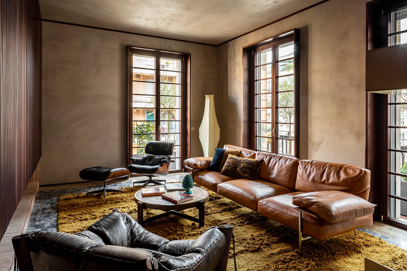 A cozy living room with a brown leather sofa, armchairs, a round coffee table, and large windows letting in natural light.