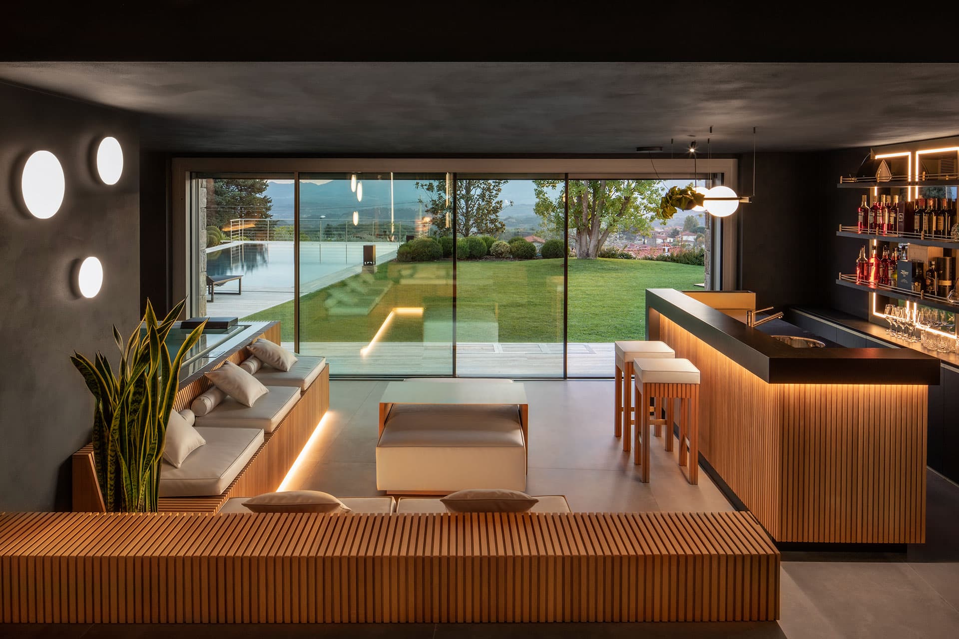 Modern lounge with wooden furniture, a sleek bar, and large windows overlooking a garden. Circular wall lights add a stylish touch.