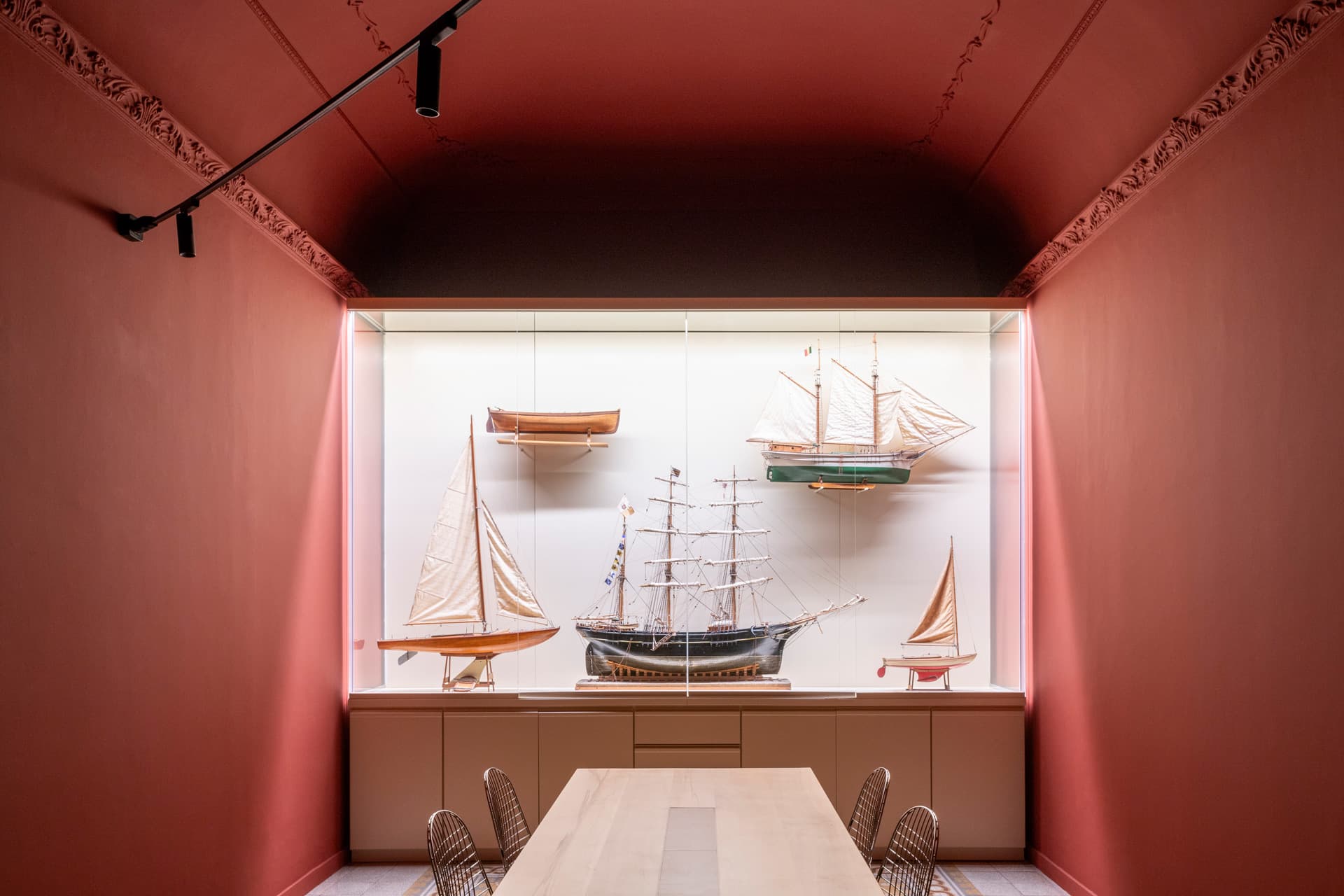 Room with red walls featuring a display case of model ships. A wooden table and wire chairs are in the foreground.