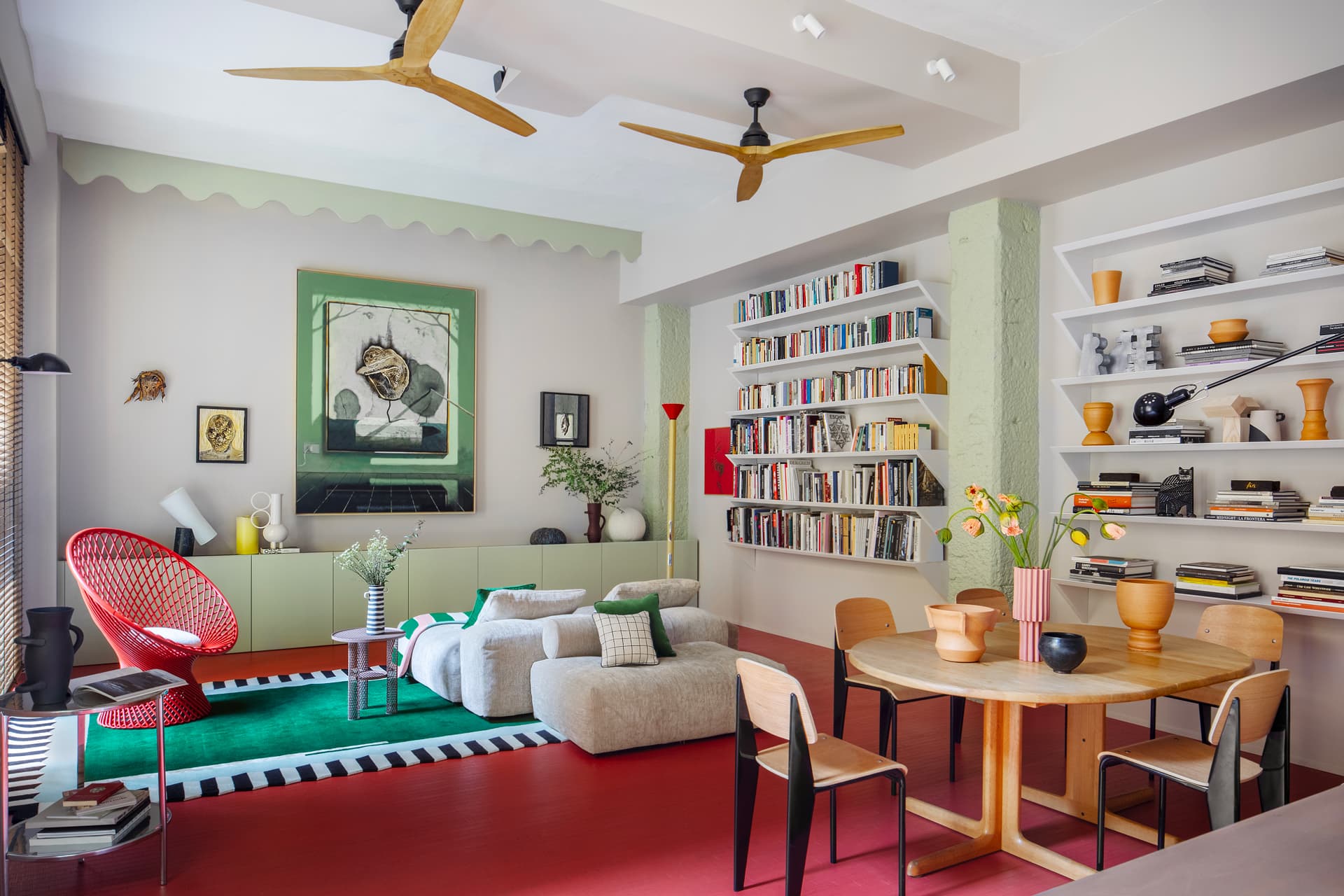 Colorful living room with red floor, modern furniture, bookshelves, and art. Features a round dining wooden table, red chair, and green accents.