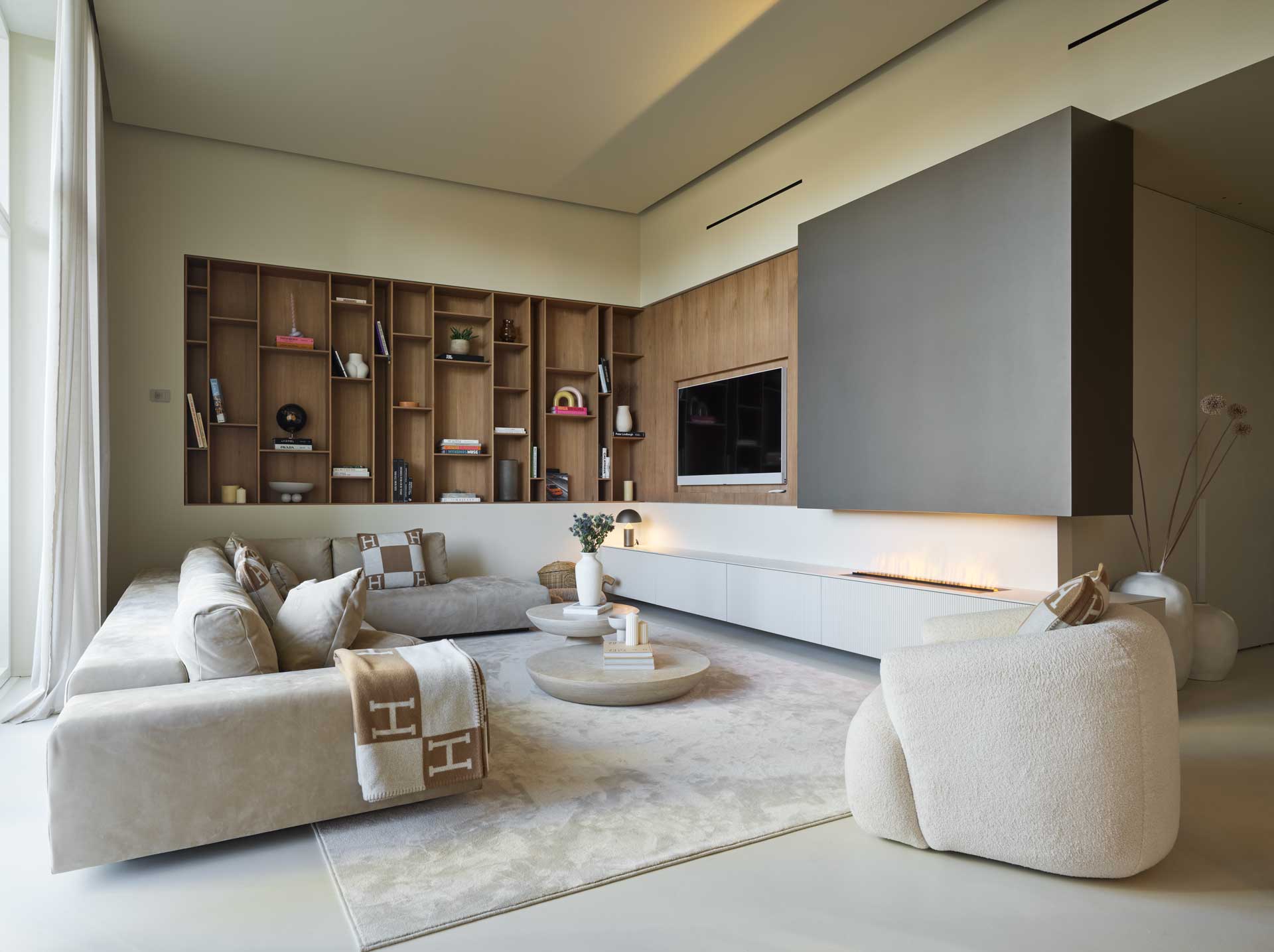 Modern living room with beige sofas, a large rug, wall-mounted TV, built-in shelves, and a sleek fireplace. Soft lighting and neutral tones.