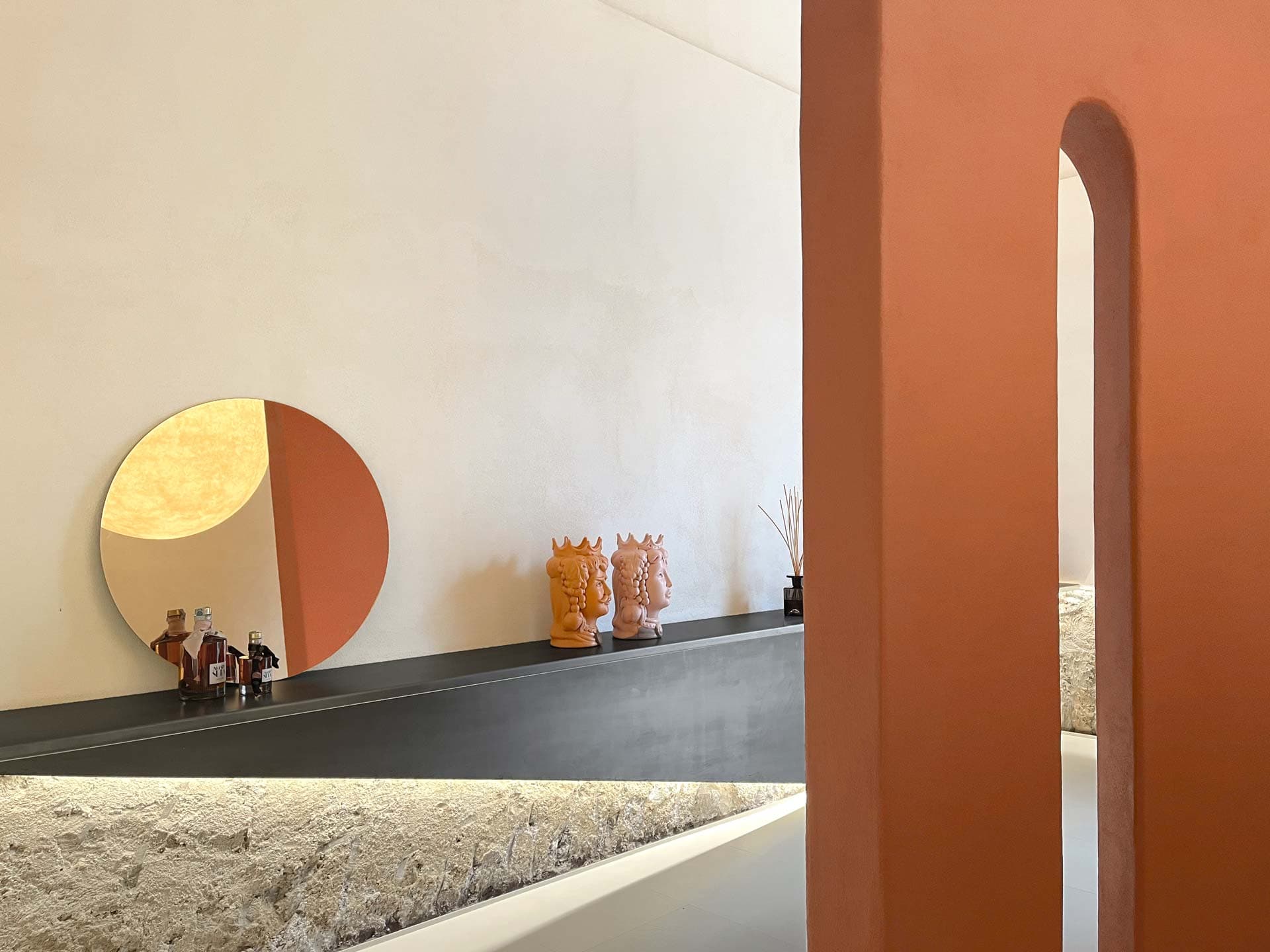 Minimalist interior with a round mirror, orange decorative masks, and a sleek shelf against a textured wall. Orange and white color scheme.