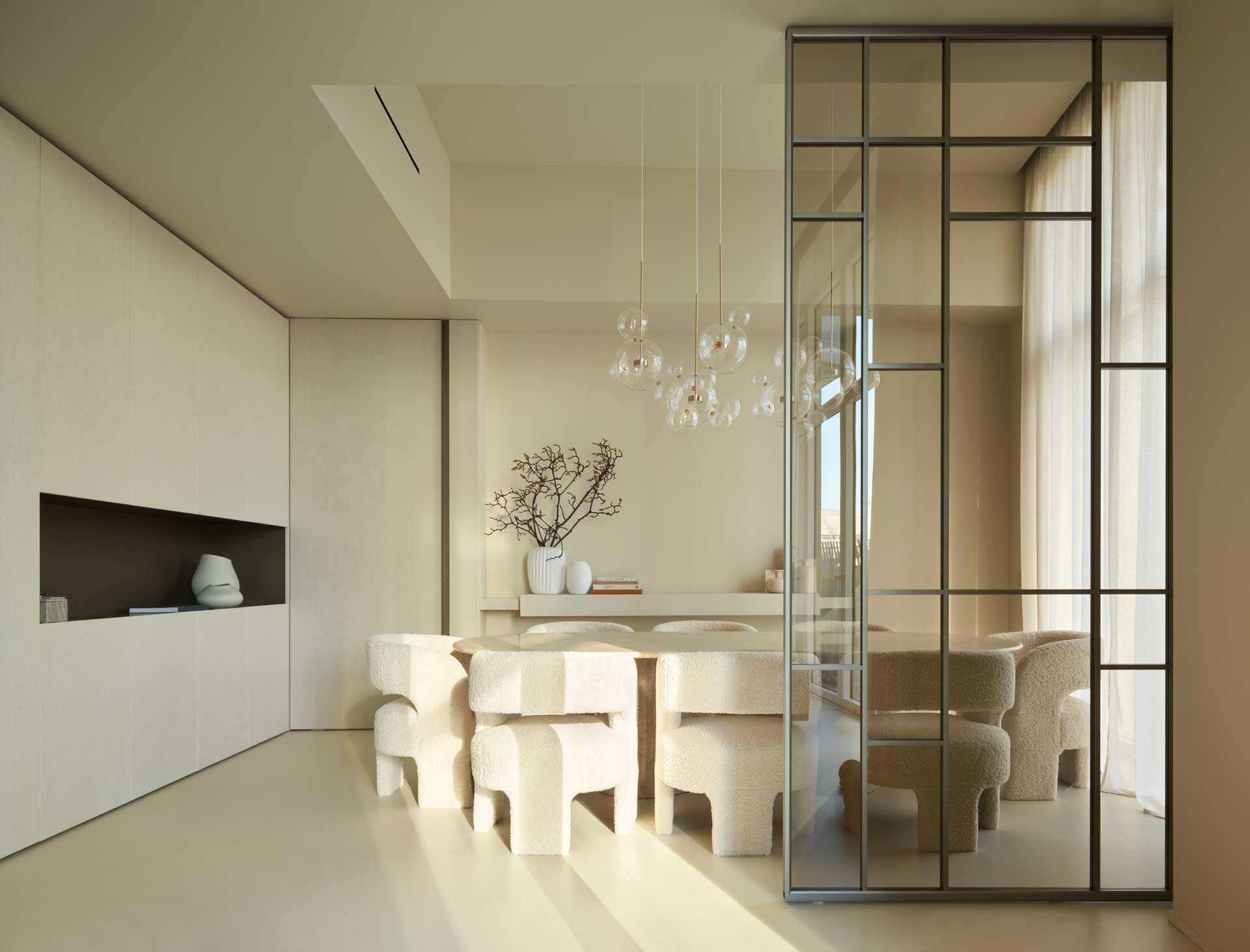Minimalist dining room with beige tones, featuring a large table, unique chairs, glass pendant lights, and a glass partition.