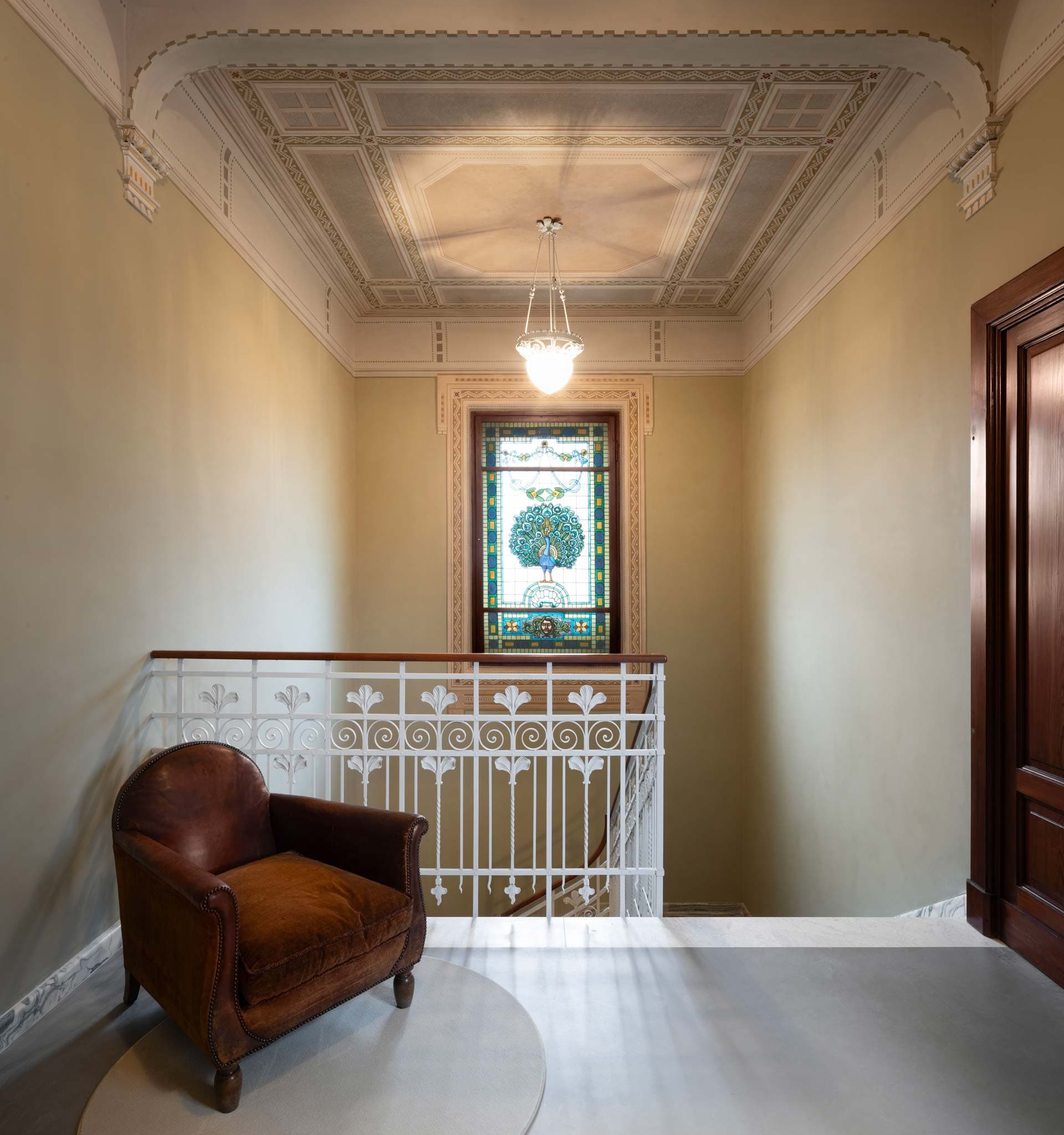 A vintage leather chair sits by a white railing in a hallway, with a decorative stained glass window and ornate ceiling above.