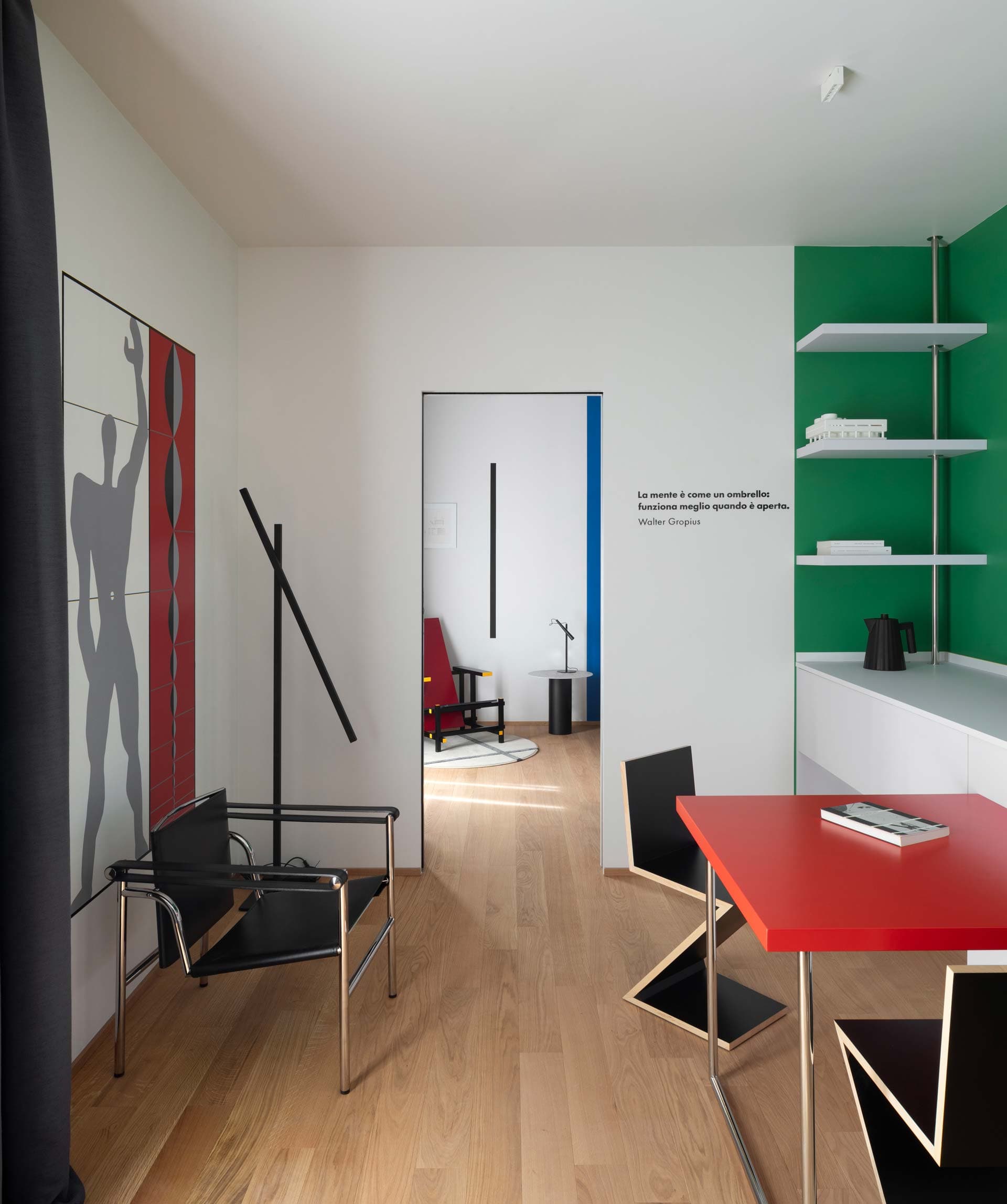 Modern interior with red table and black geometric chairs. Green accent wall, wooden floor, and open doorway to another room.