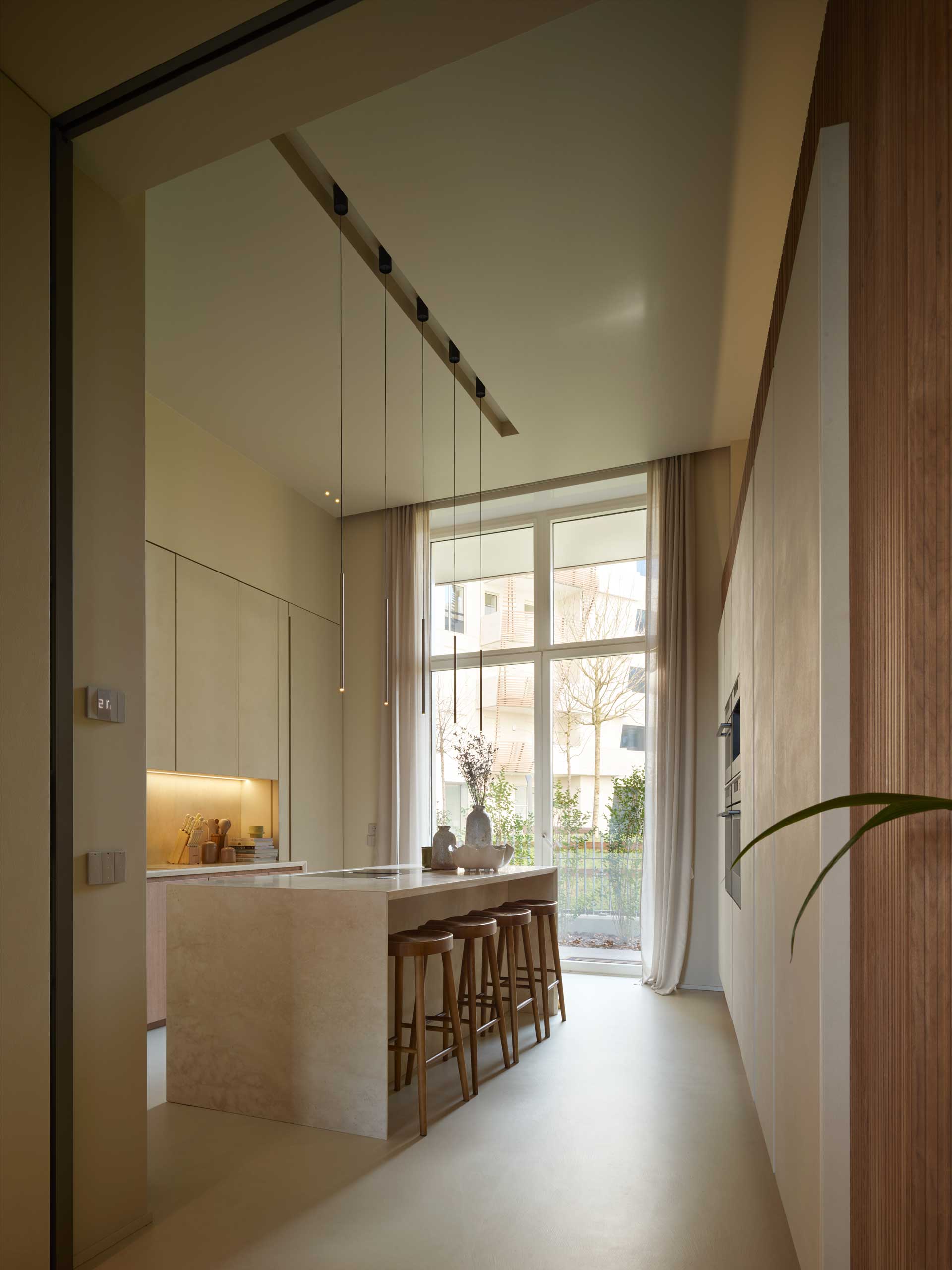 Modern kitchen with high ceiling, large windows, marble island with wooden stools, and pendant lighting. Neutral tones create a minimalist look.