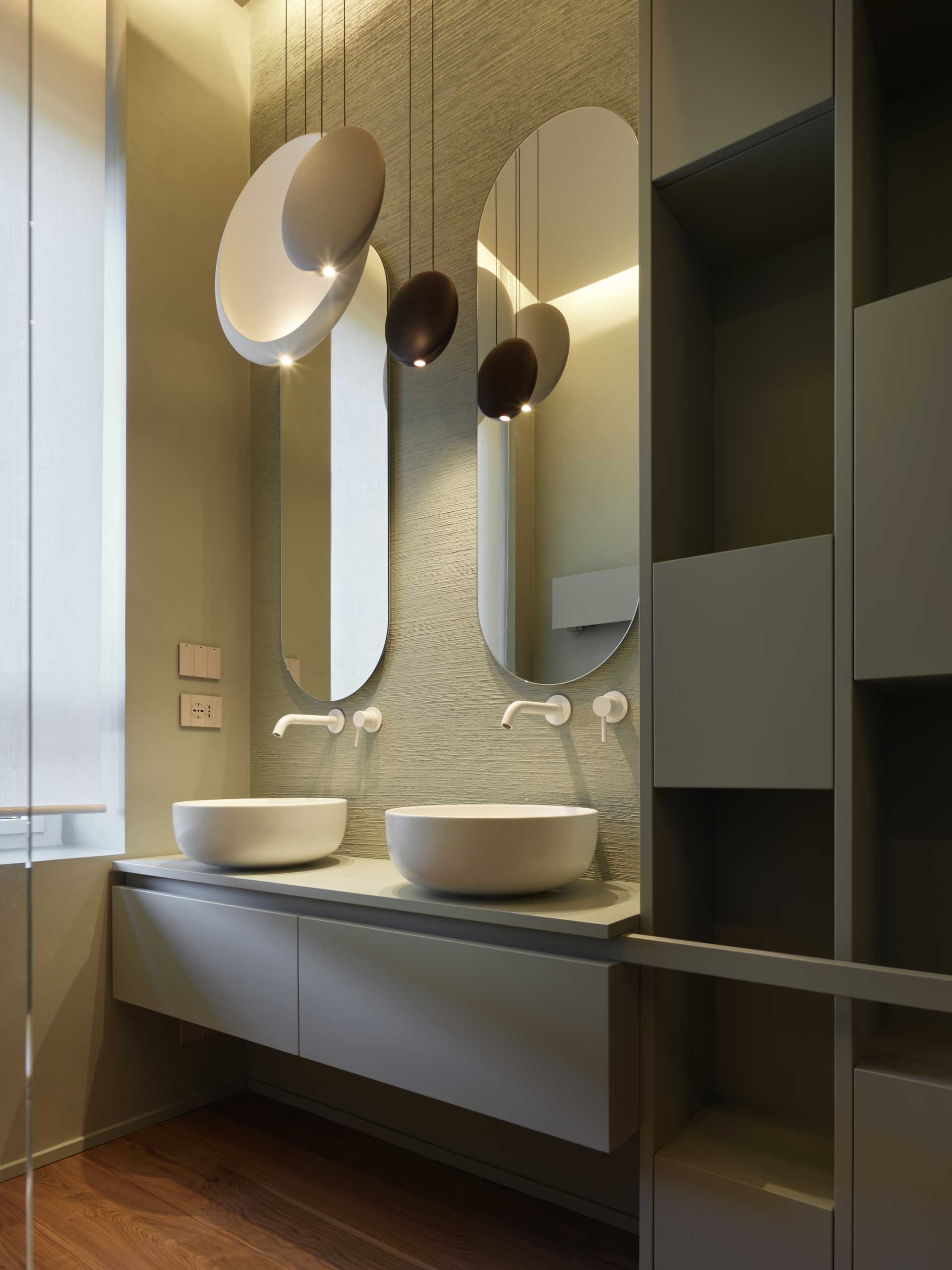 Modern bathroom with two oval mirrors, round sinks, and pendant lights. Minimalist design with neutral tones and sleek cabinetry.