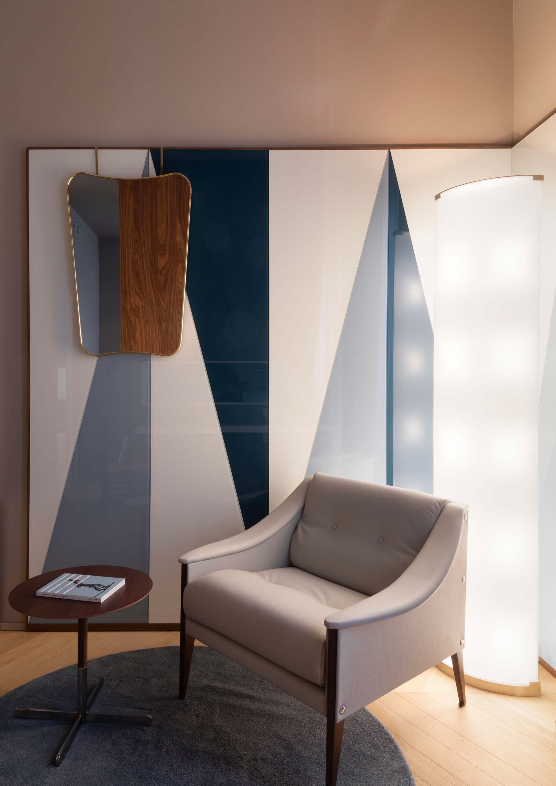 Modern room with a gray armchair, round table with a magazine, geometric wall art, and a tall, illuminated floor lamp on a wooden floor.