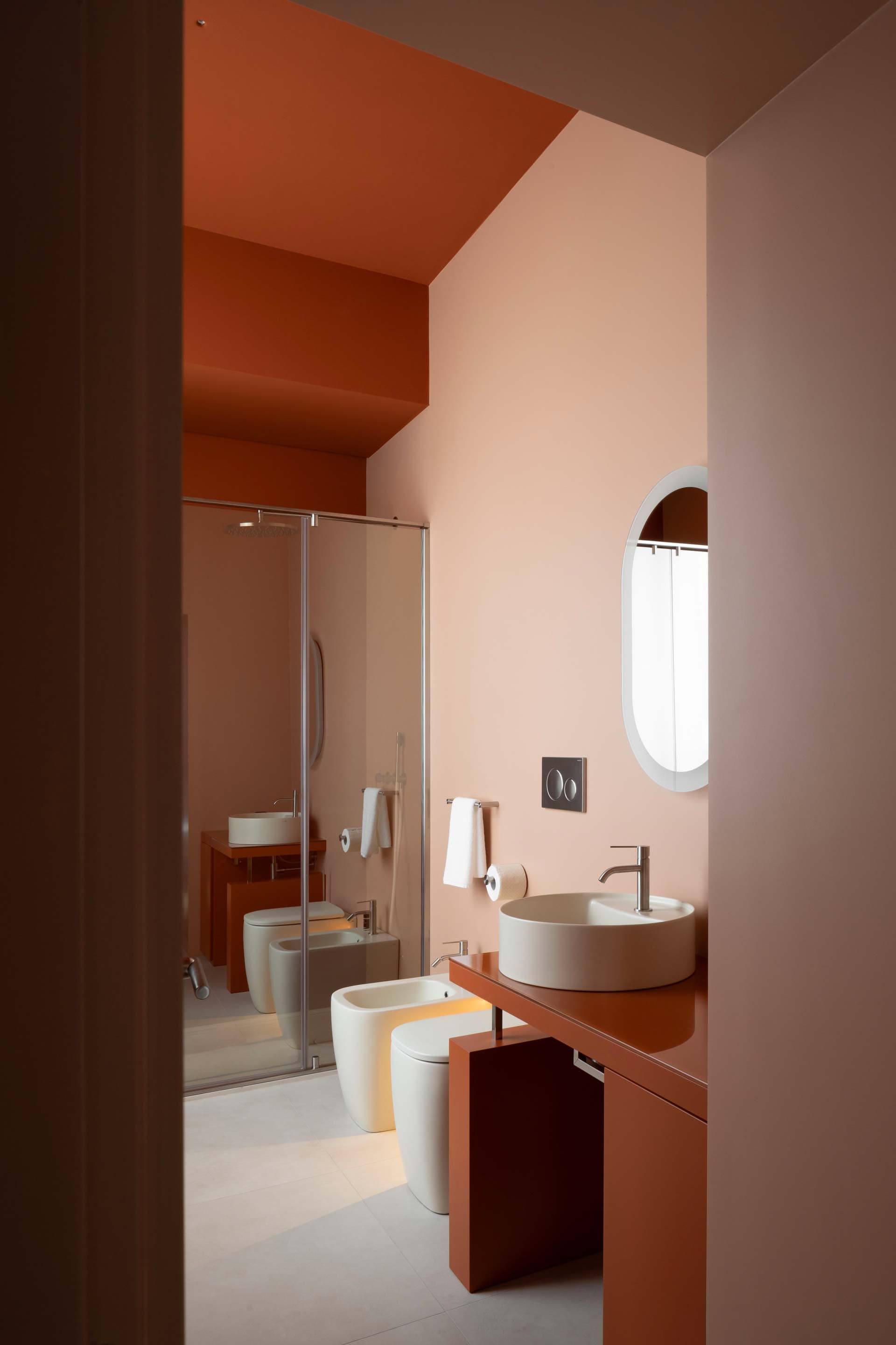 Modern bathroom with terracotta walls, round mirror, vessel sink on a counter, and glass-enclosed shower. Minimalistic and stylish design.