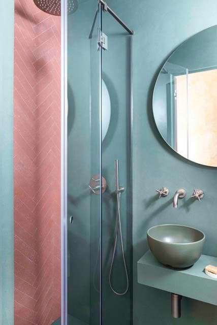Compact bathroom with a glass shower, round mirror, green wall-mounted sink, and pink herringbone tiles in the shower area.